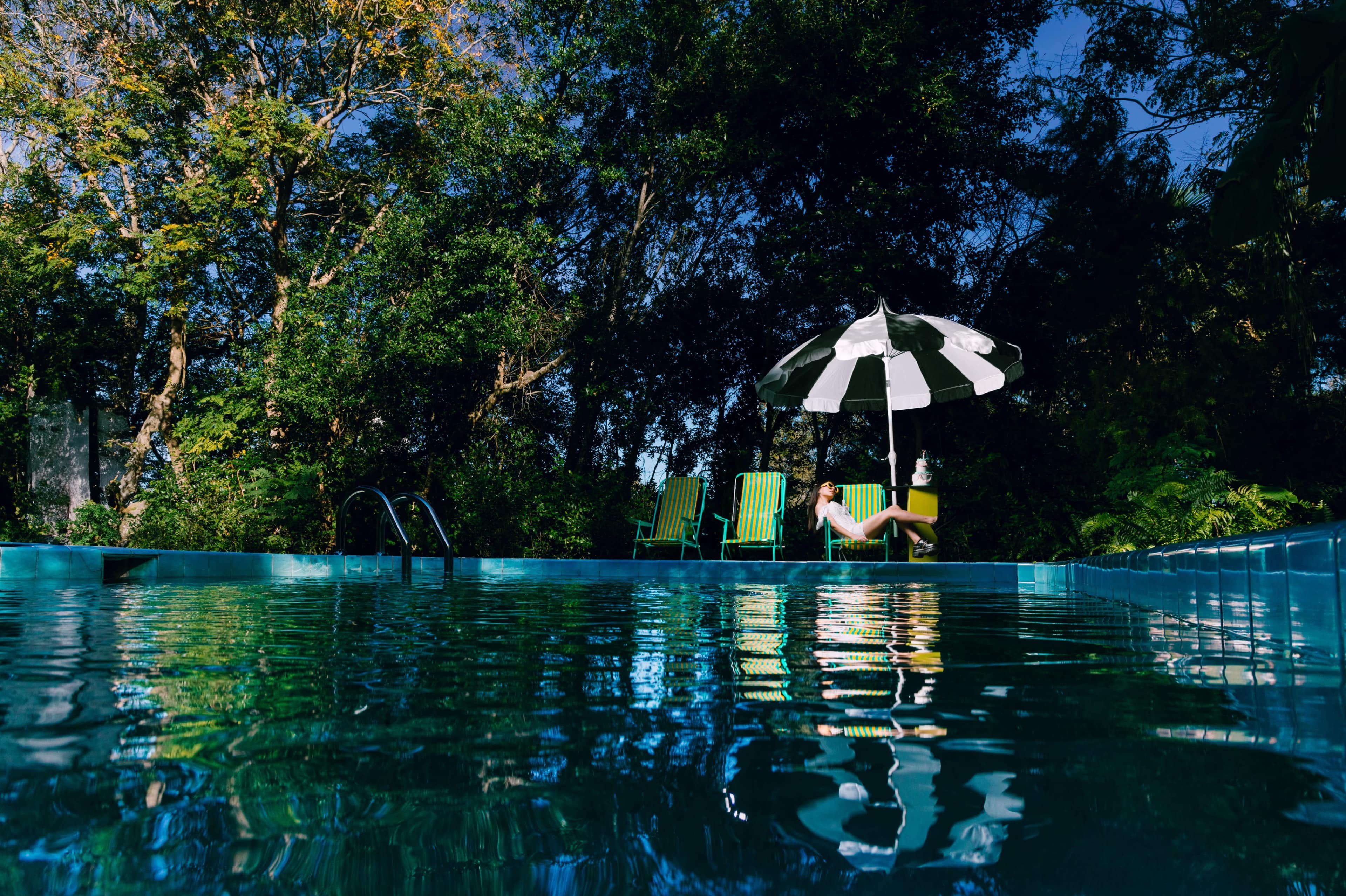 A person sits under a black-and-white umbrella beside a swimming pool surrounded by trees.