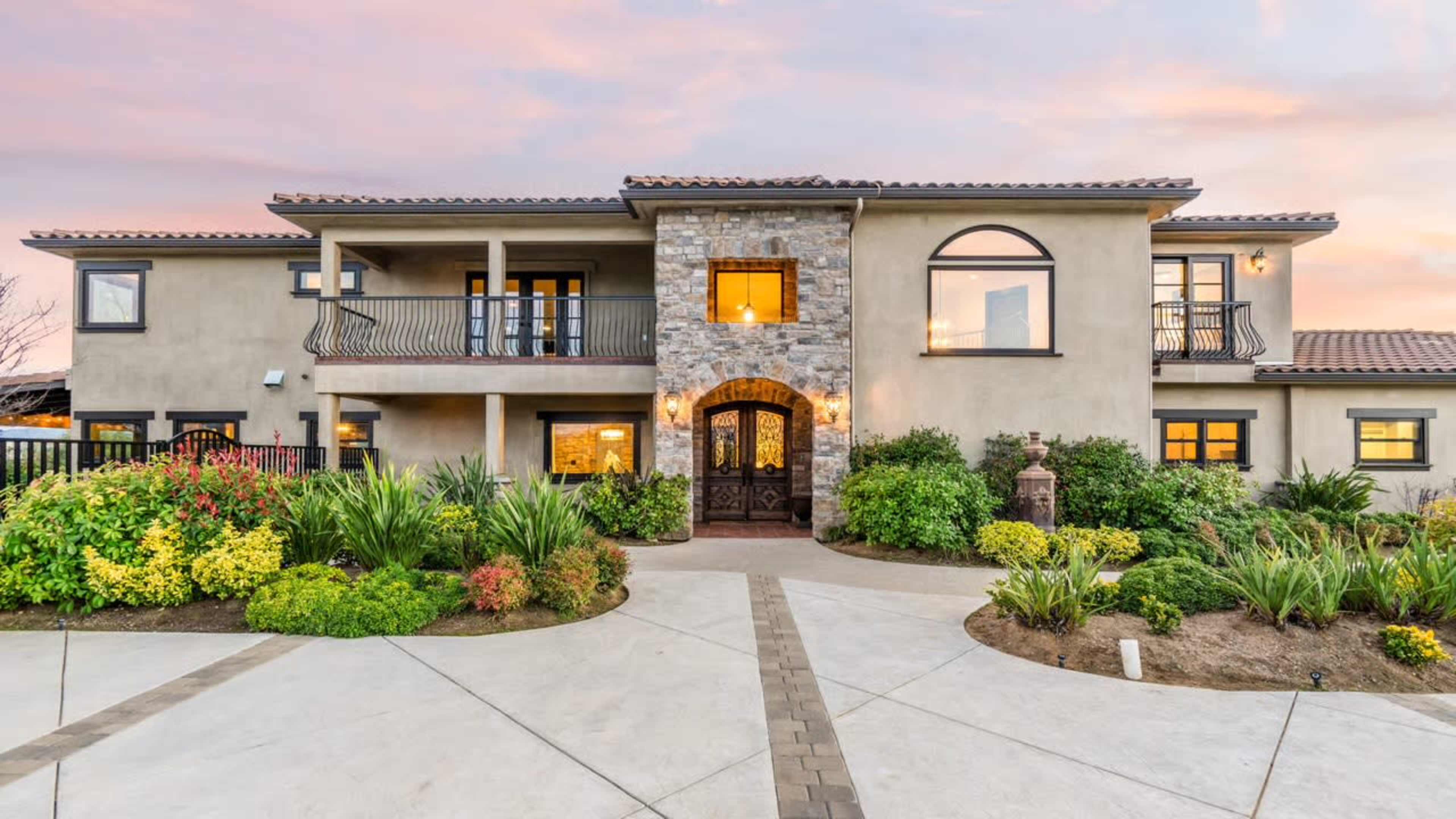 The image shows a large, modern house with a stone facade, surrounded by landscaped gardens and a paved walkway.