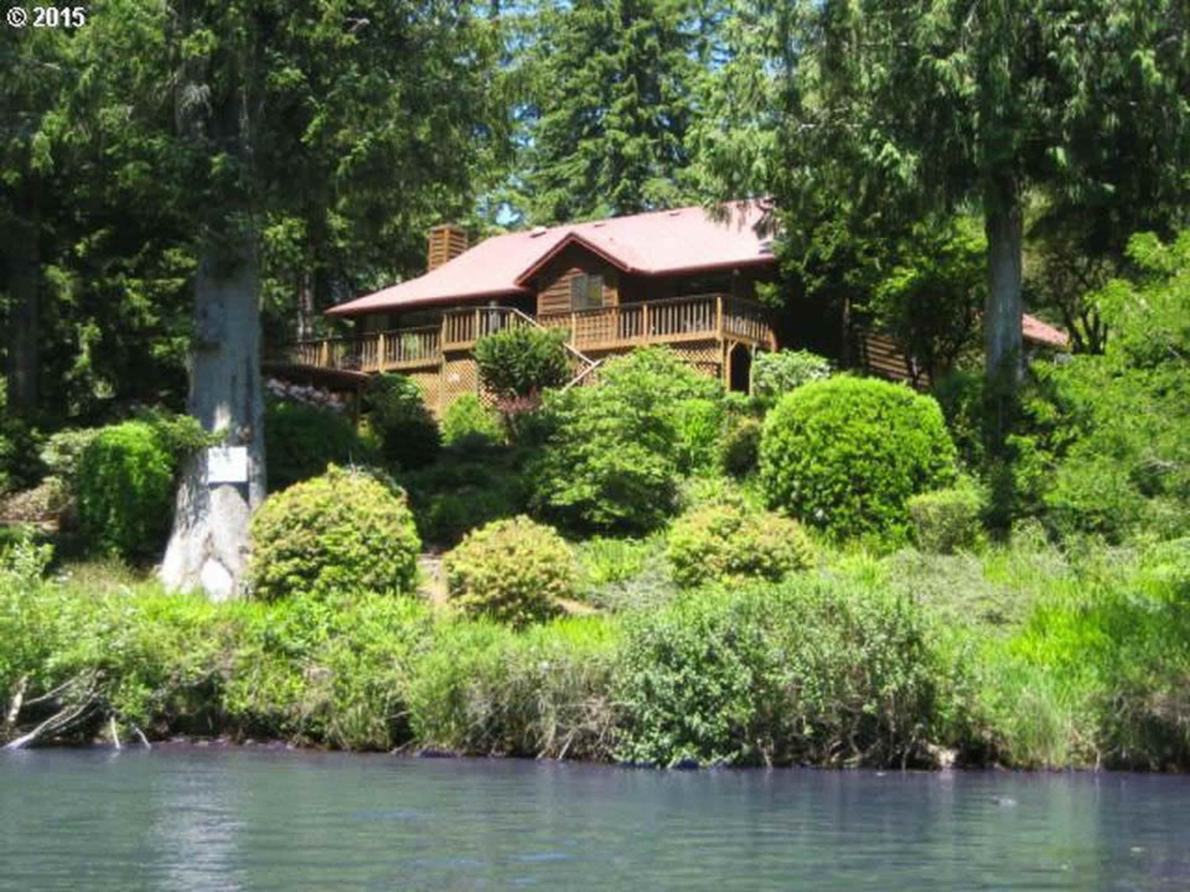 A large wooden house with a red roof is situated on a hillside surrounded by dense greenery and trees.