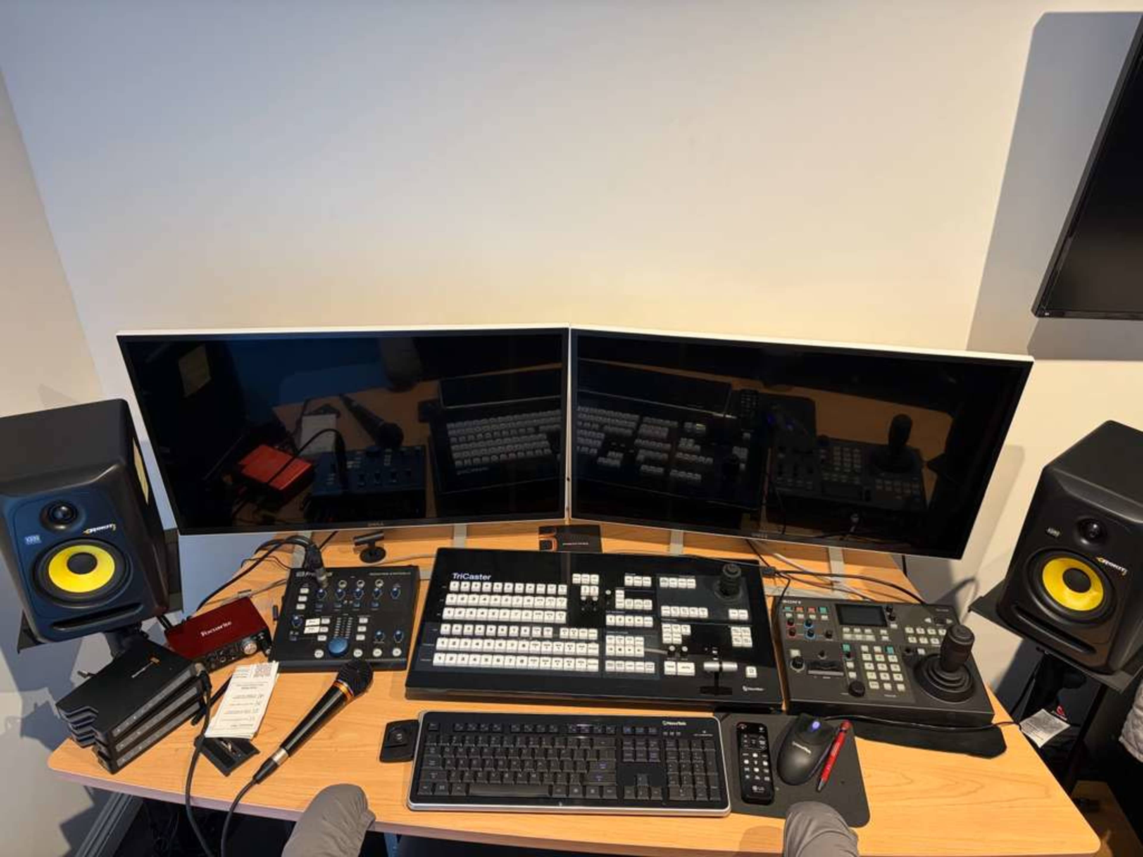 The image shows a digital editing workstation featuring two large monitors, a control panel with buttons and dials, a keyboard, speakers, and various tools organized on a wooden desk.