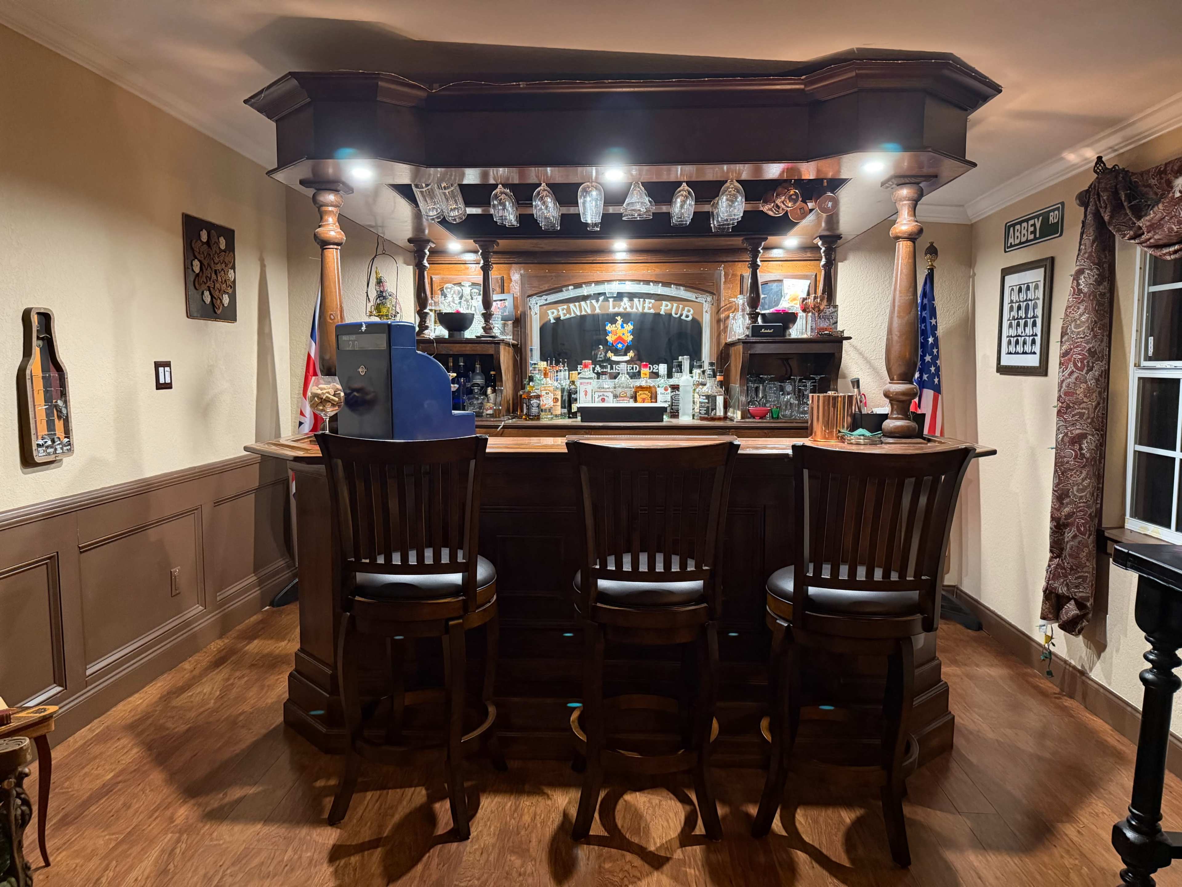 The image shows a wooden home bar with three high-backed chairs, shelves lined with glassware, and various decorations, including flags and artwork on the walls.