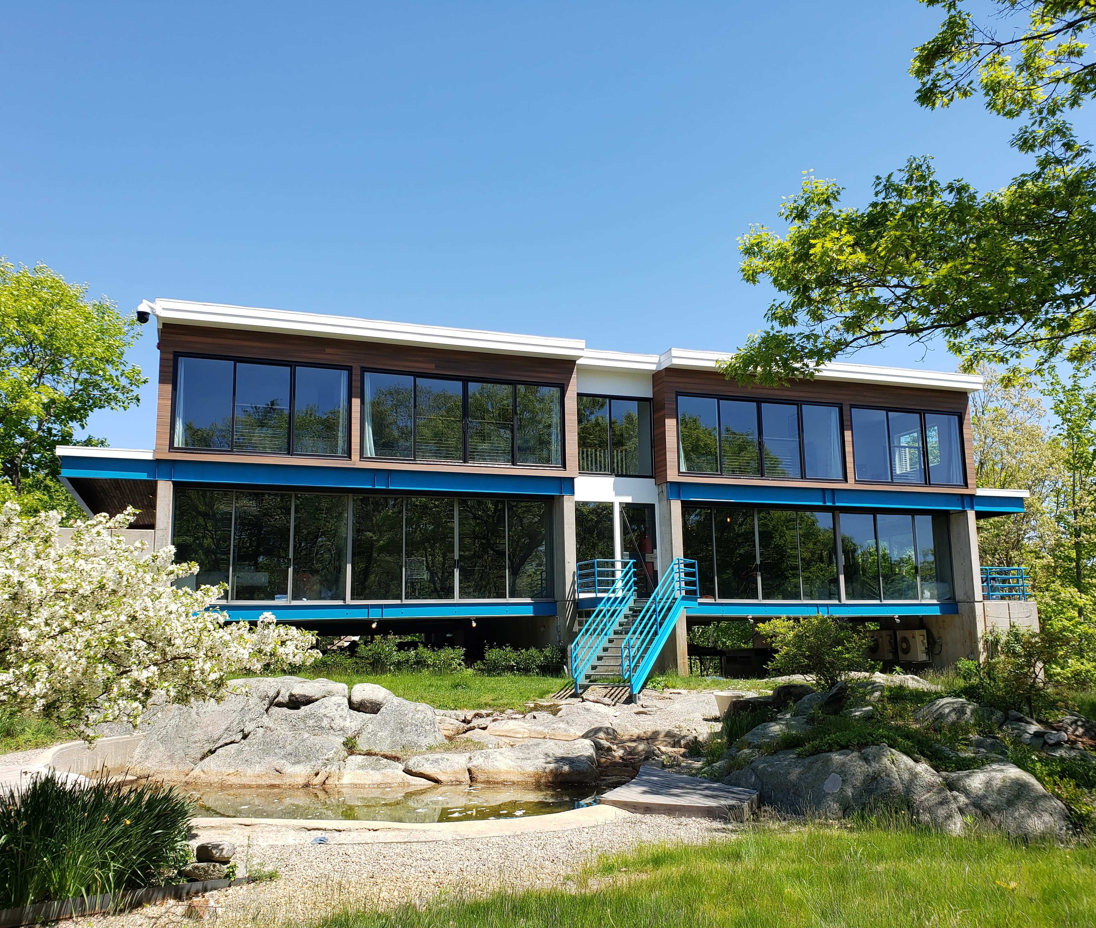 A modern, two-story house with large glass windows and a blue staircase is set amidst greenery and boulders.