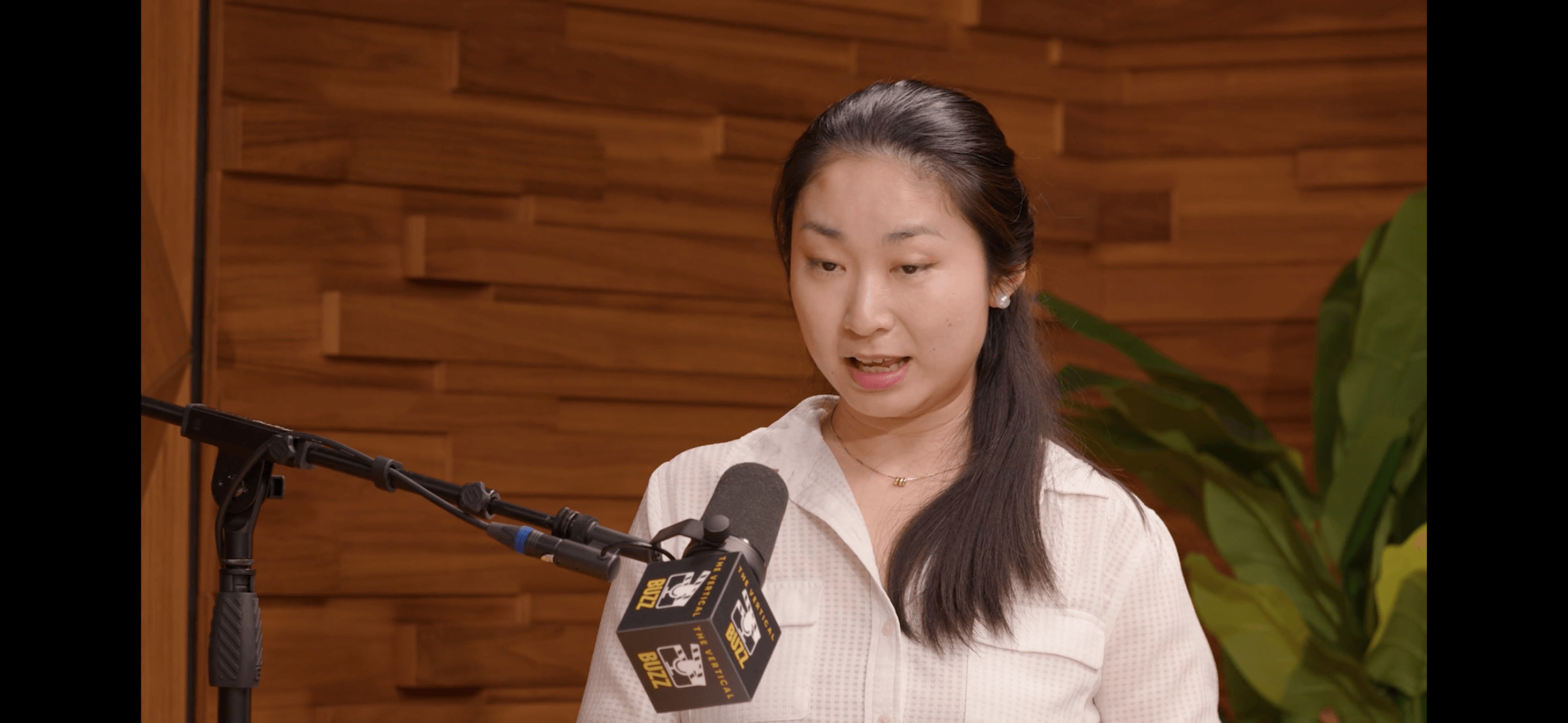A woman speaks into a microphone while standing in front of a wooden backdrop with plants.