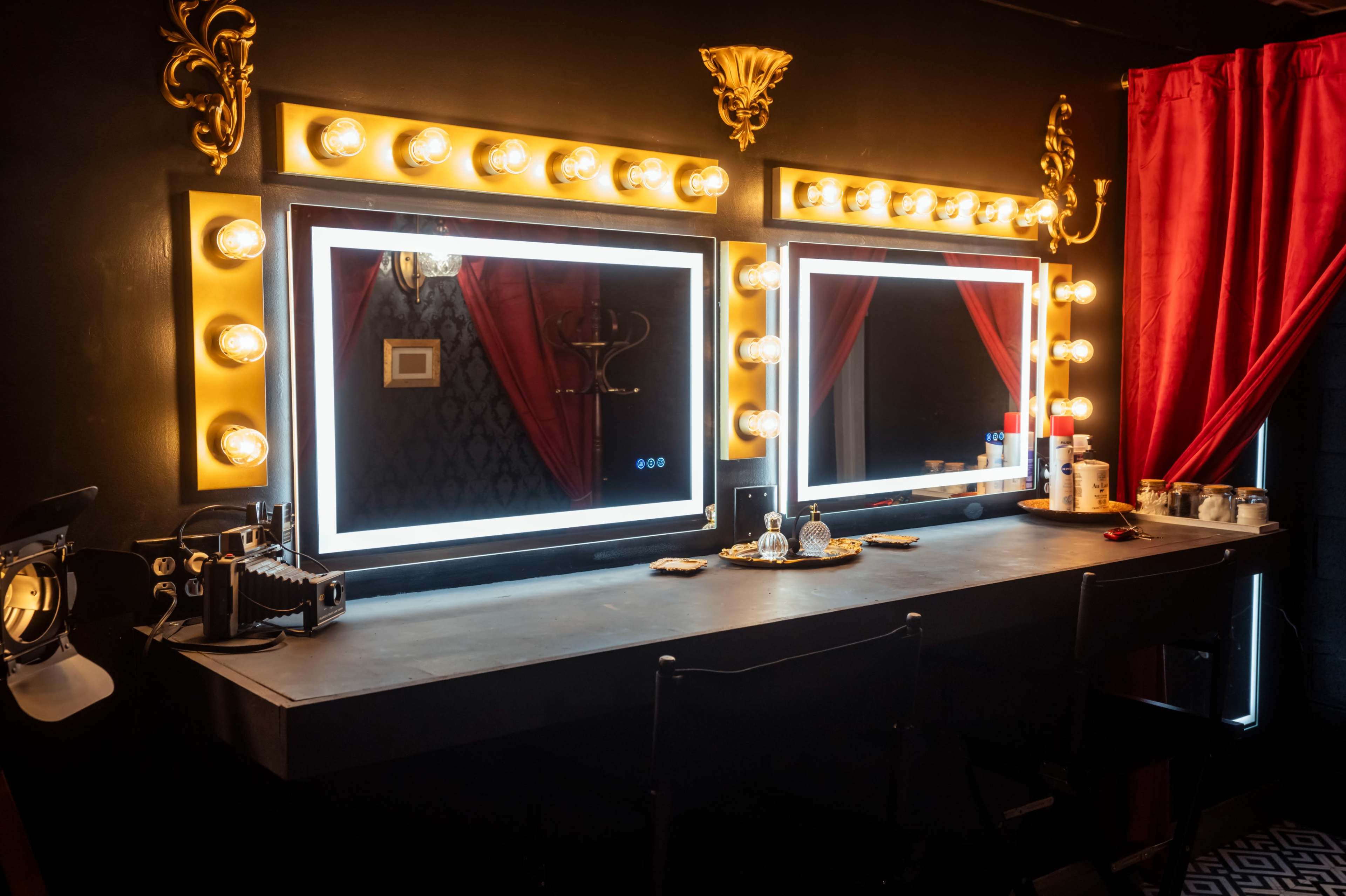 A dimly lit dressing room with two illuminated mirrors surrounded by light bulbs, a long table with various cosmetic items, and red curtains in the background.
