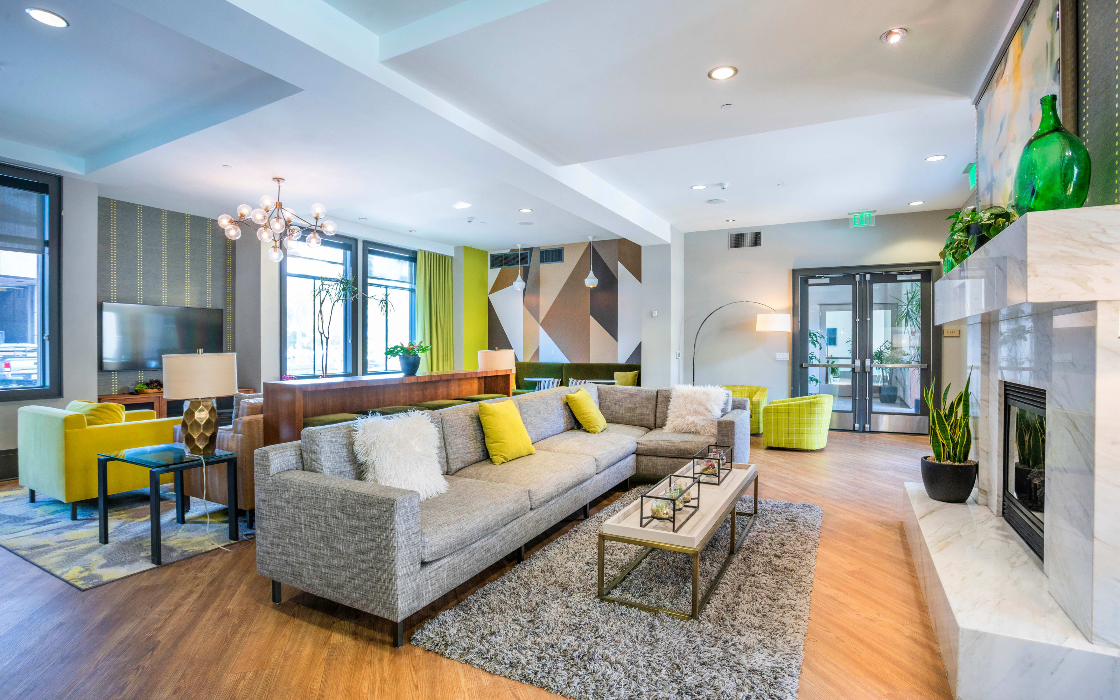 A modern lounge area featuring a gray sectional sofa, a coffee table, a fireplace, and various seating options in a brightly lit space with decorative plants and artwork.