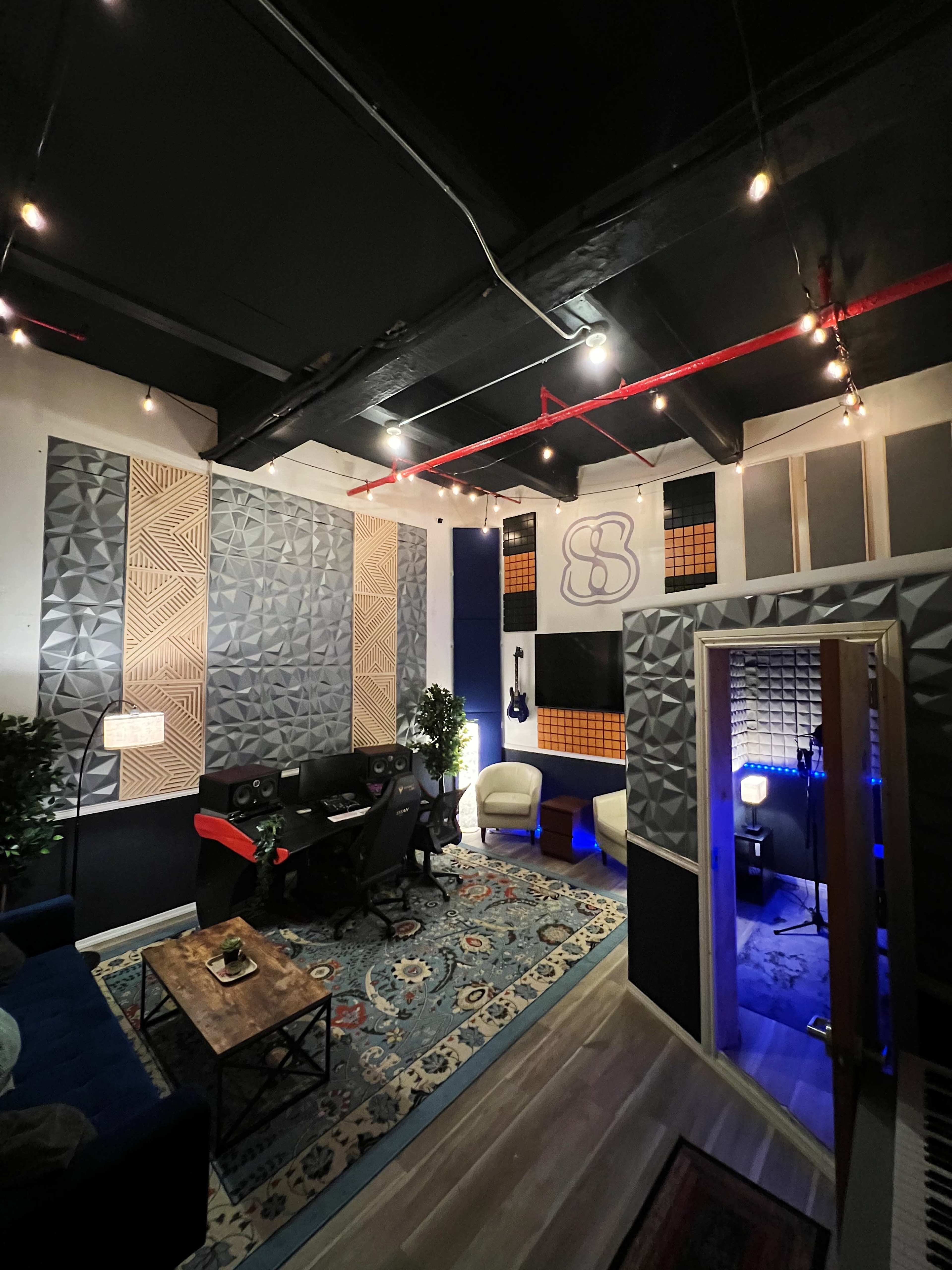 A music recording studio features soundproof walls, a control desk with computers, and cozy seating areas, illuminated by string lights.