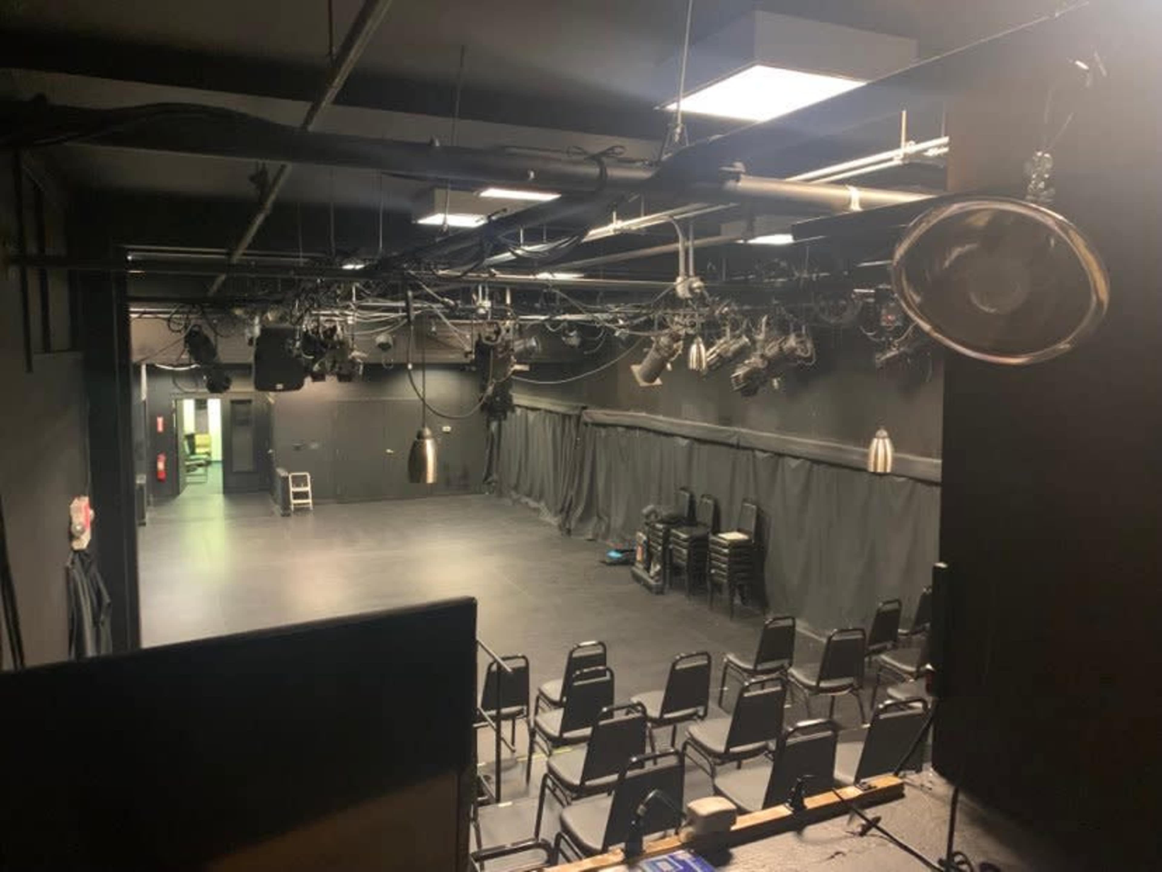 The image shows a small black-box theater with empty chairs arranged in rows and stage lighting overhead.