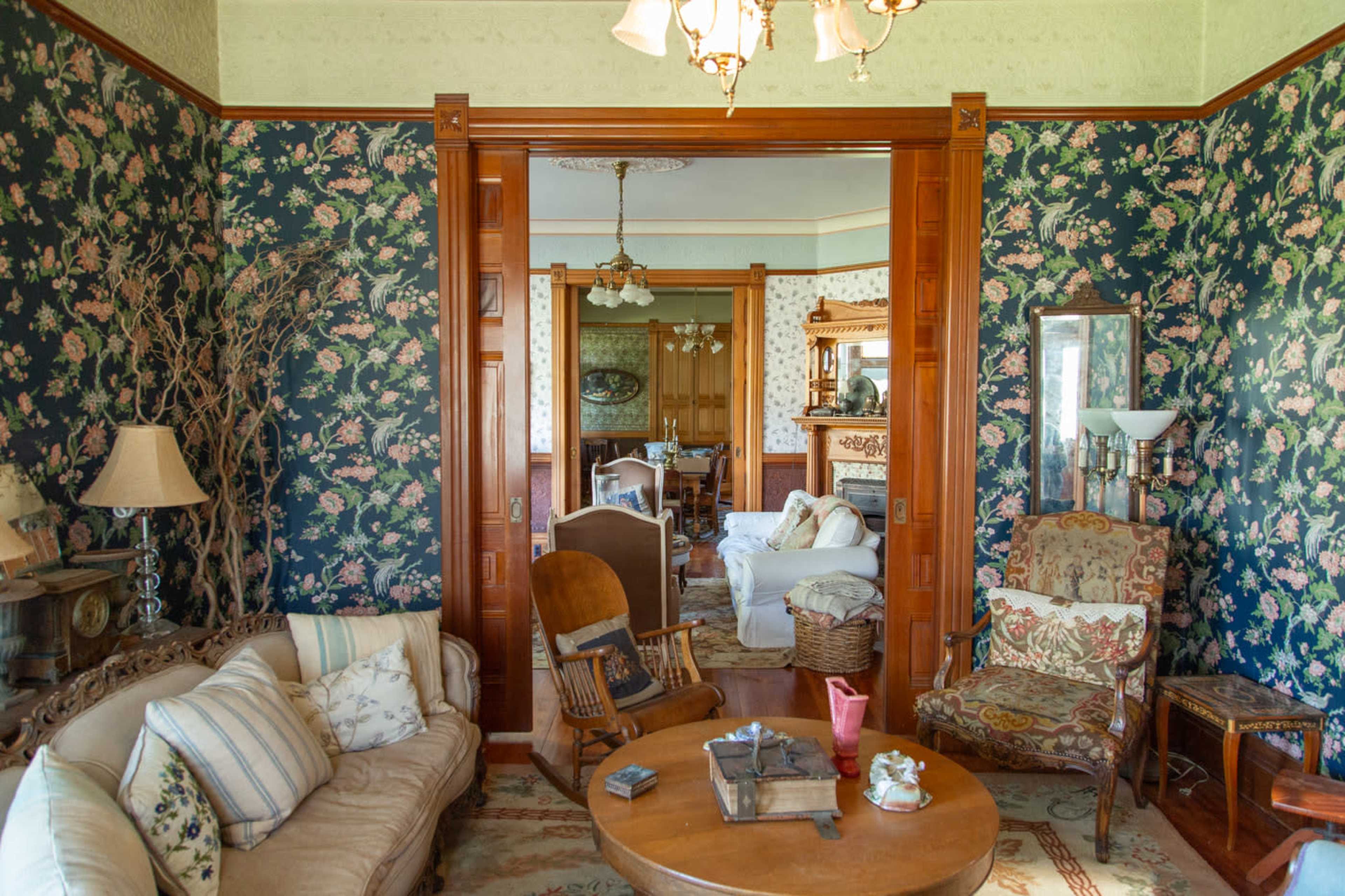 The image depicts a vintage living room with floral wallpaper, wooden furniture, and a view into a connected room featuring a fireplace and additional seating.