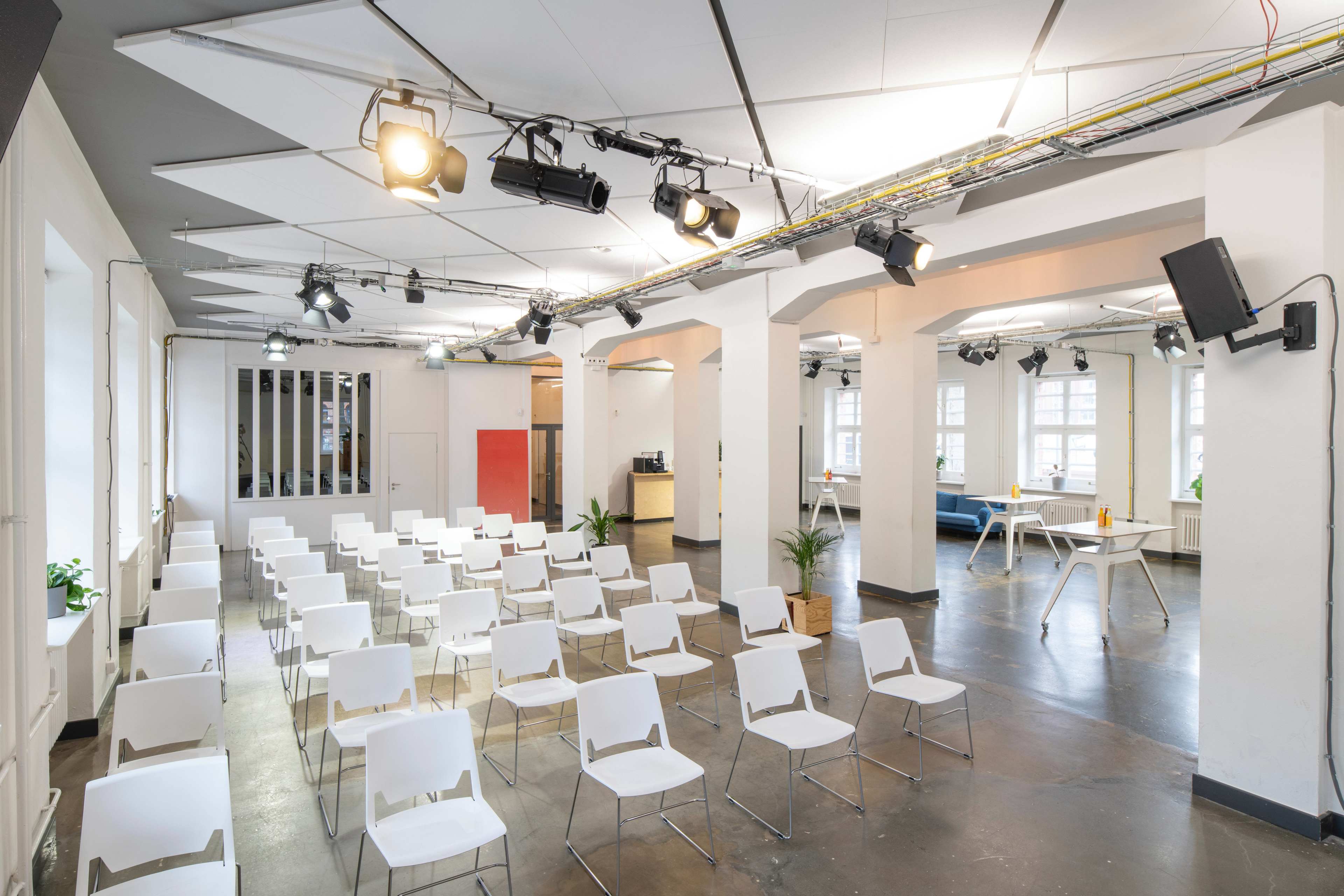 A spacious seminar room arranged with white chairs facing a central area, illuminated by overhead lighting and featuring large windows.