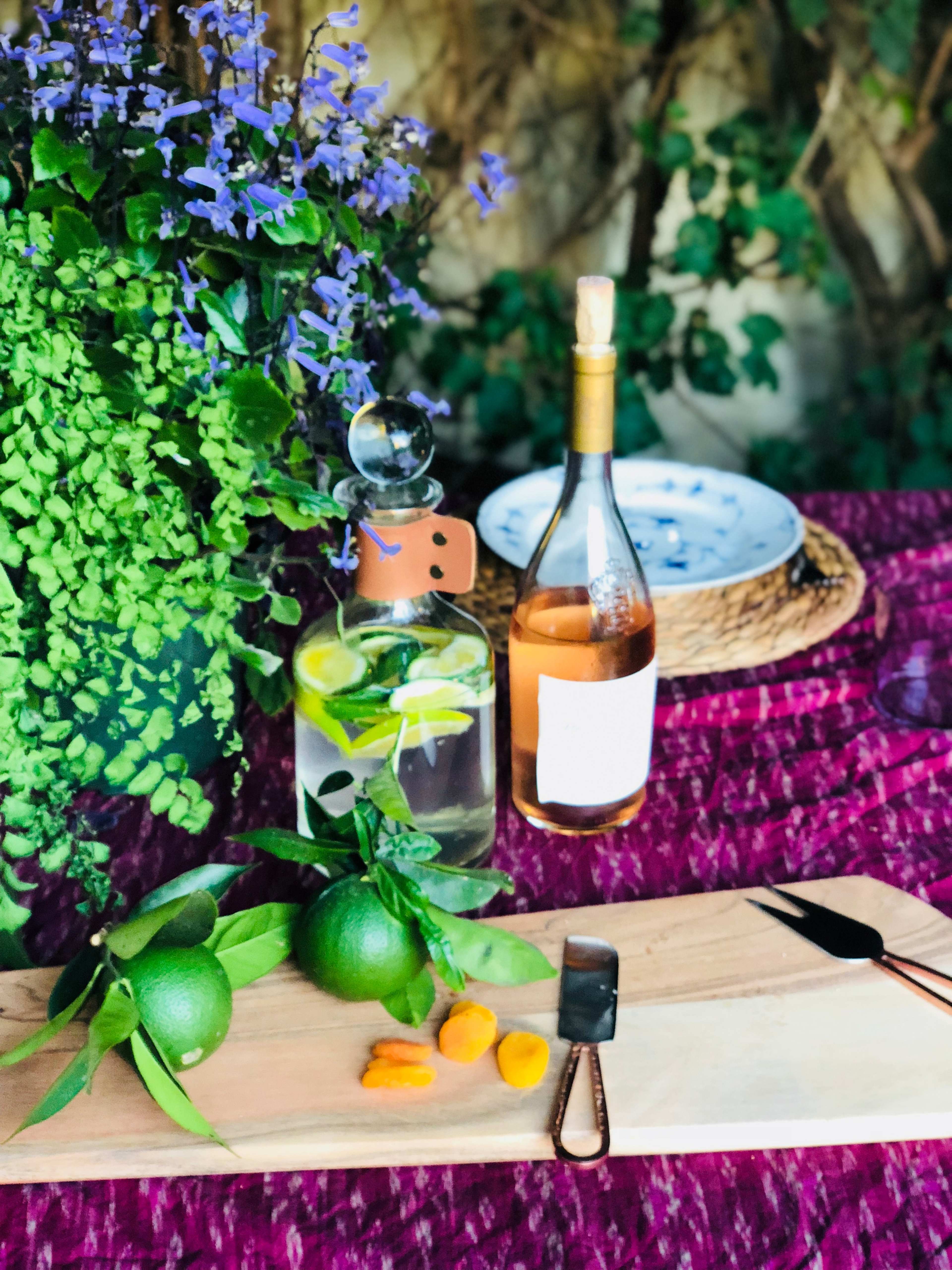 A wooden serving board with limes, a knife, and a bottle of rosé wine, surrounded by a vibrant arrangement of greenery and flowers.