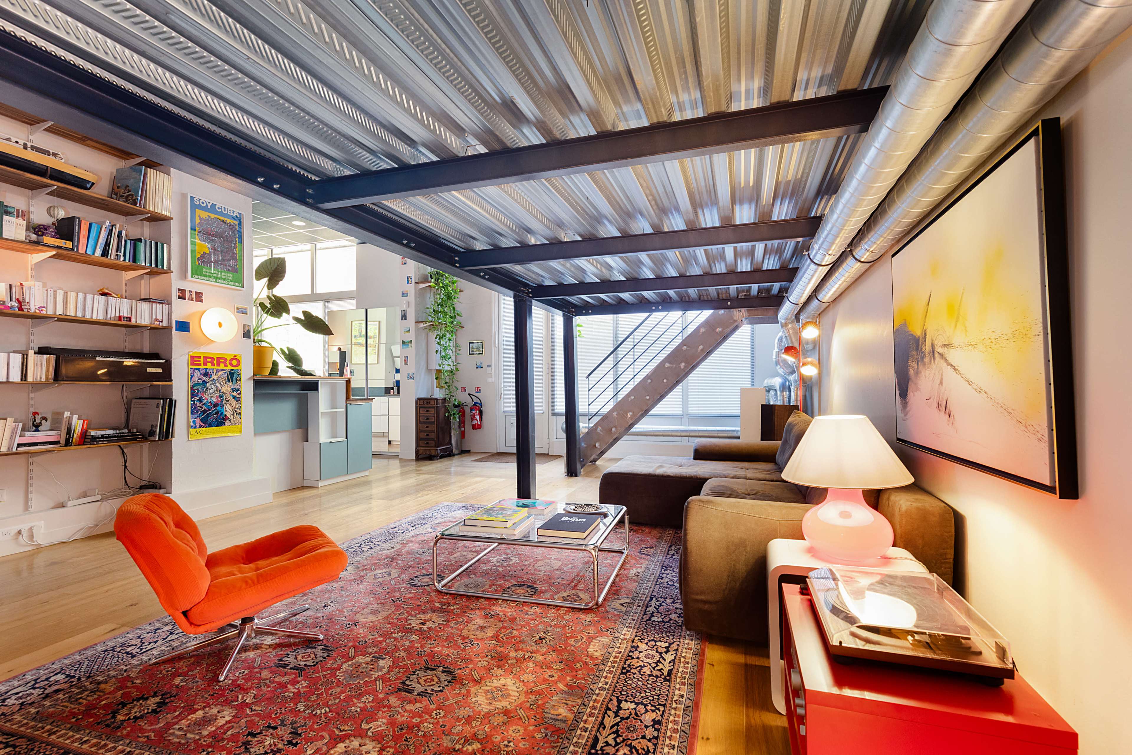 The image shows a modern living area with a sofa, an orange chair, a coffee table on a patterned rug, and a staircase leading to a loft space.