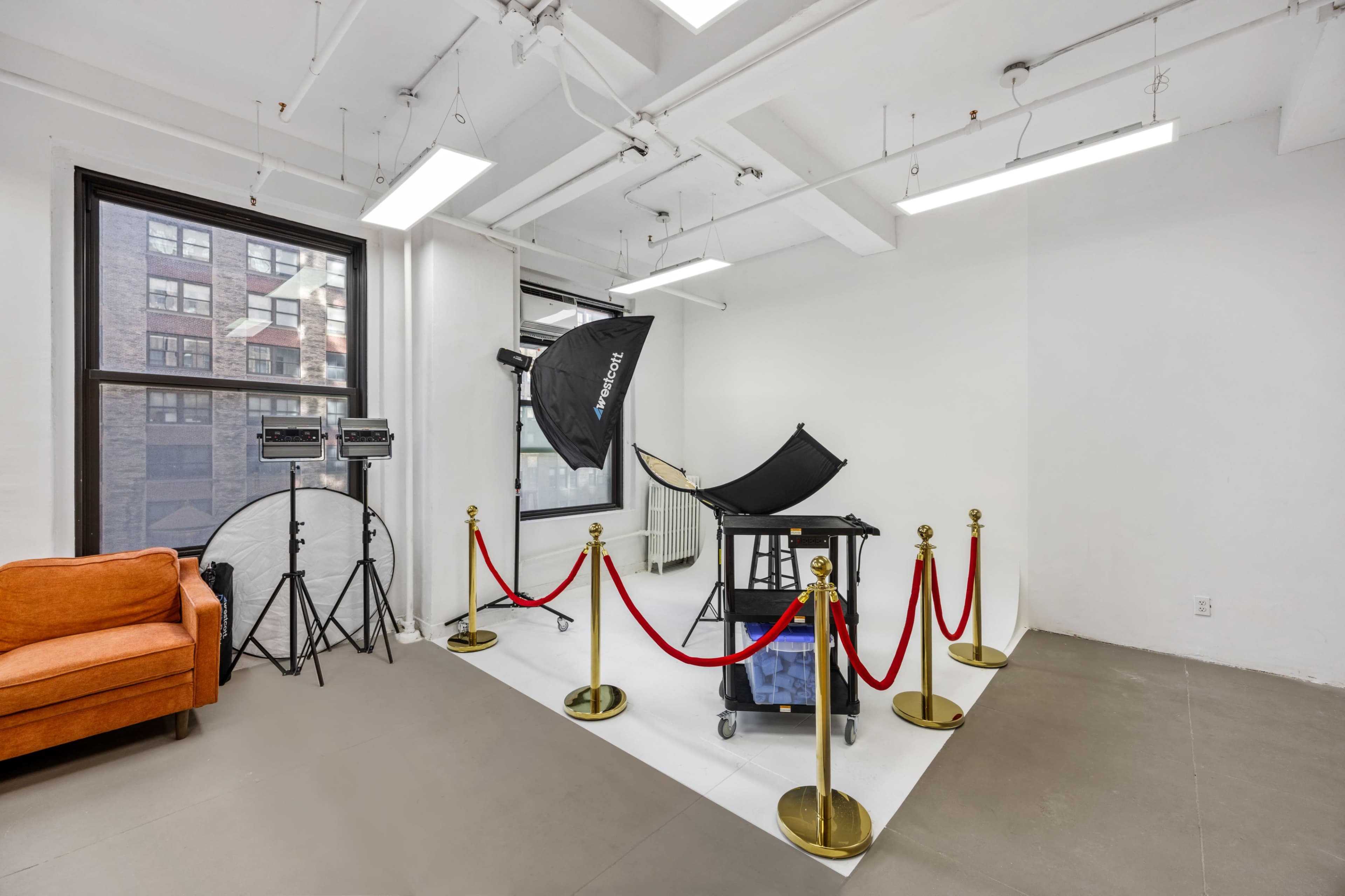The image shows a studio space with a photo setup, including lighting equipment, a backdrop, and a velvet rope barrier, alongside an orange couch against a wall of windows.