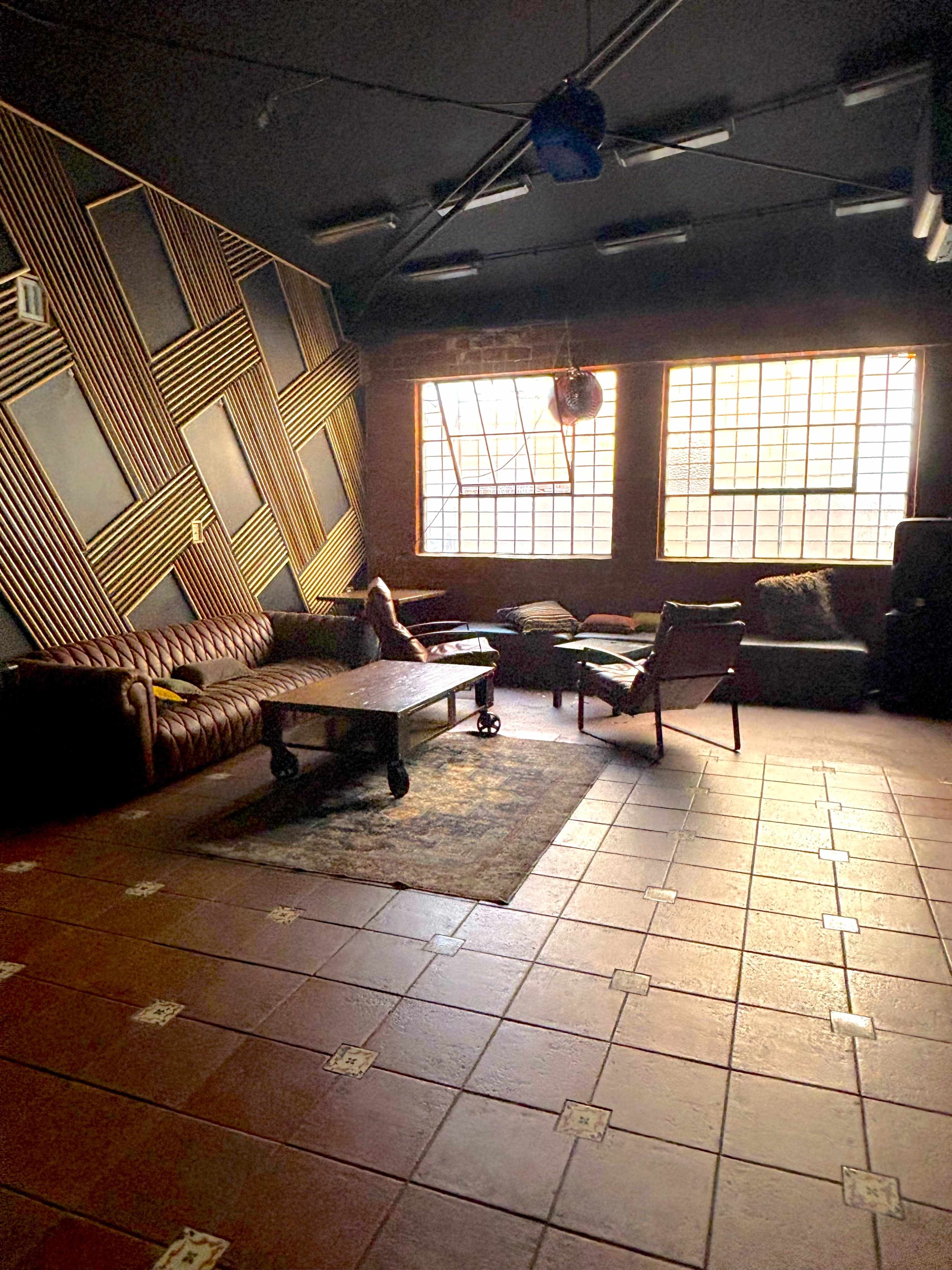 The image shows a dimly lit lounge area with a leather sofa, a wooden coffee table, and several armchairs, complemented by large windows that allow some natural light.