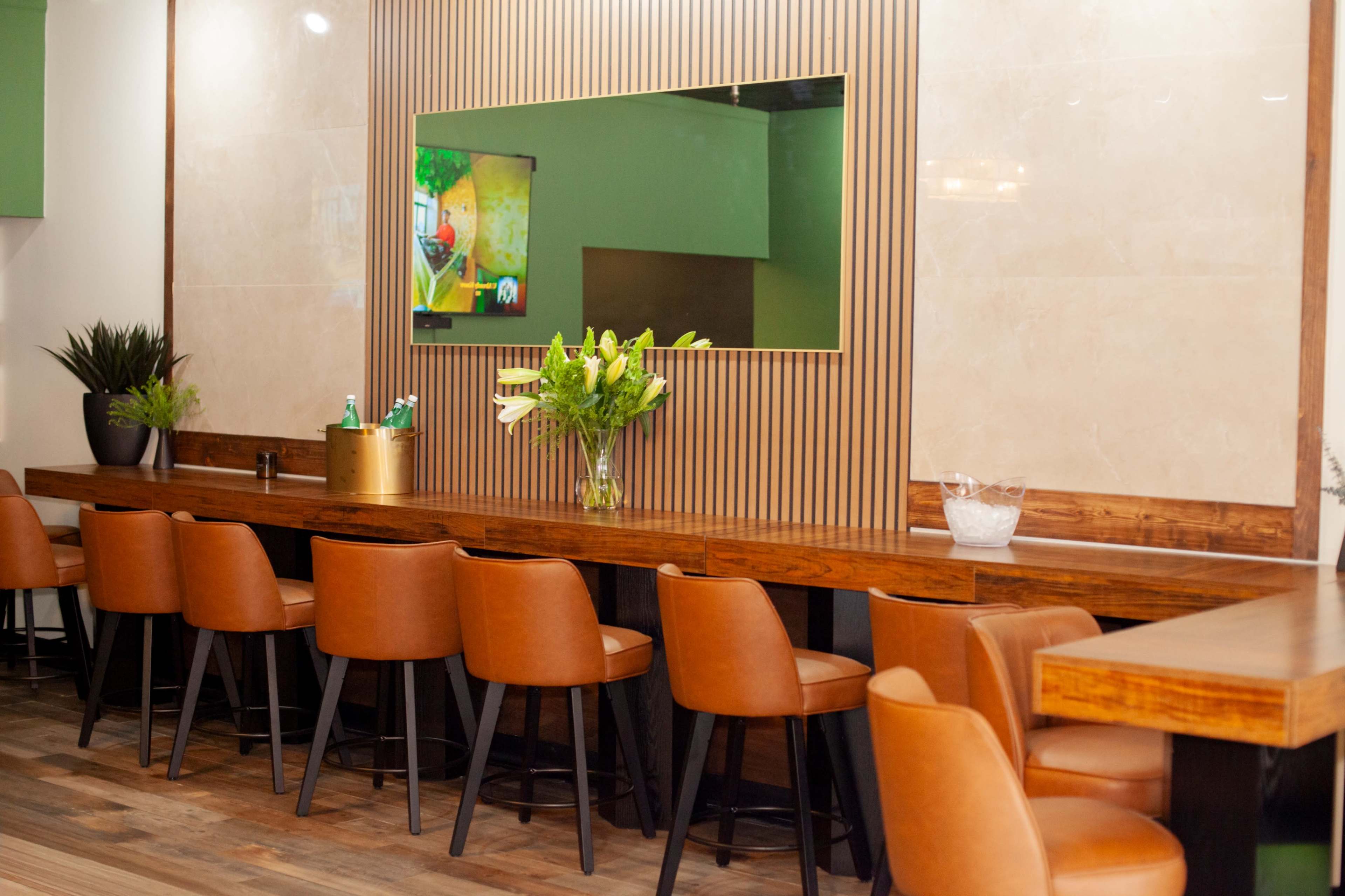 The image shows a modern interior of a restaurant featuring a wooden bar with brown leather stools, a large mirror, and a television on the wall.