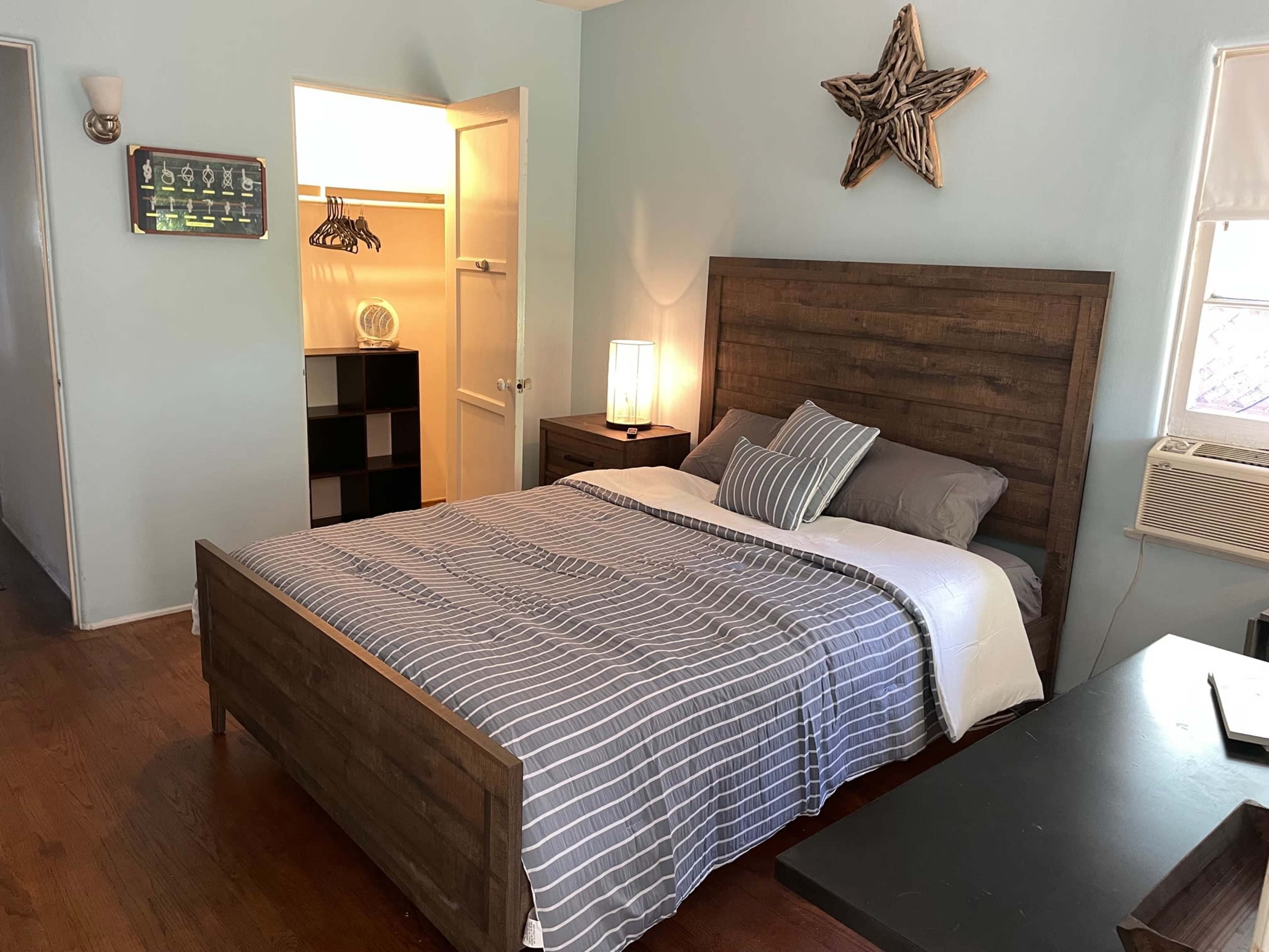 A neatly arranged bedroom features a wooden bed with gray-striped bedding, a nightstand with a lamp, and a closet with an open door revealing storage space.