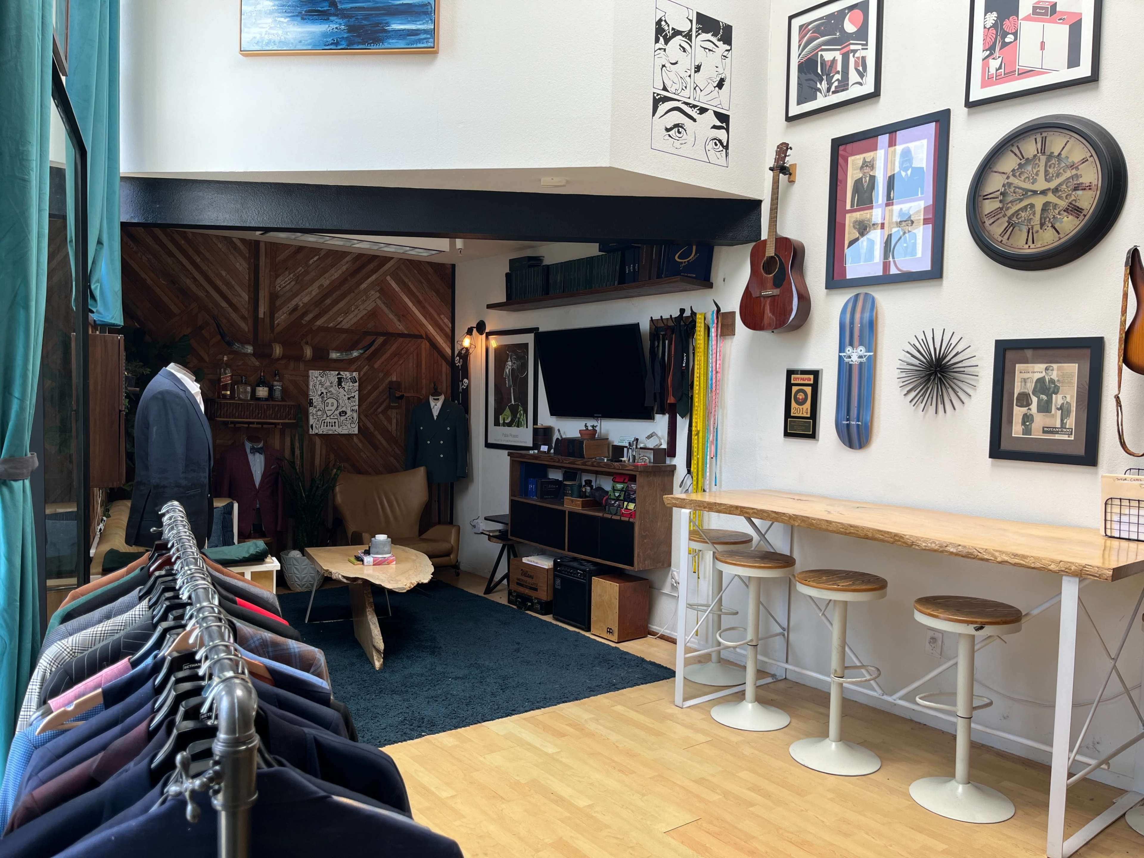 A boutique interior featuring colorful clothing racks on the left, a cozy seating area with a television and art on the walls, and a wooden table with bar stools.
