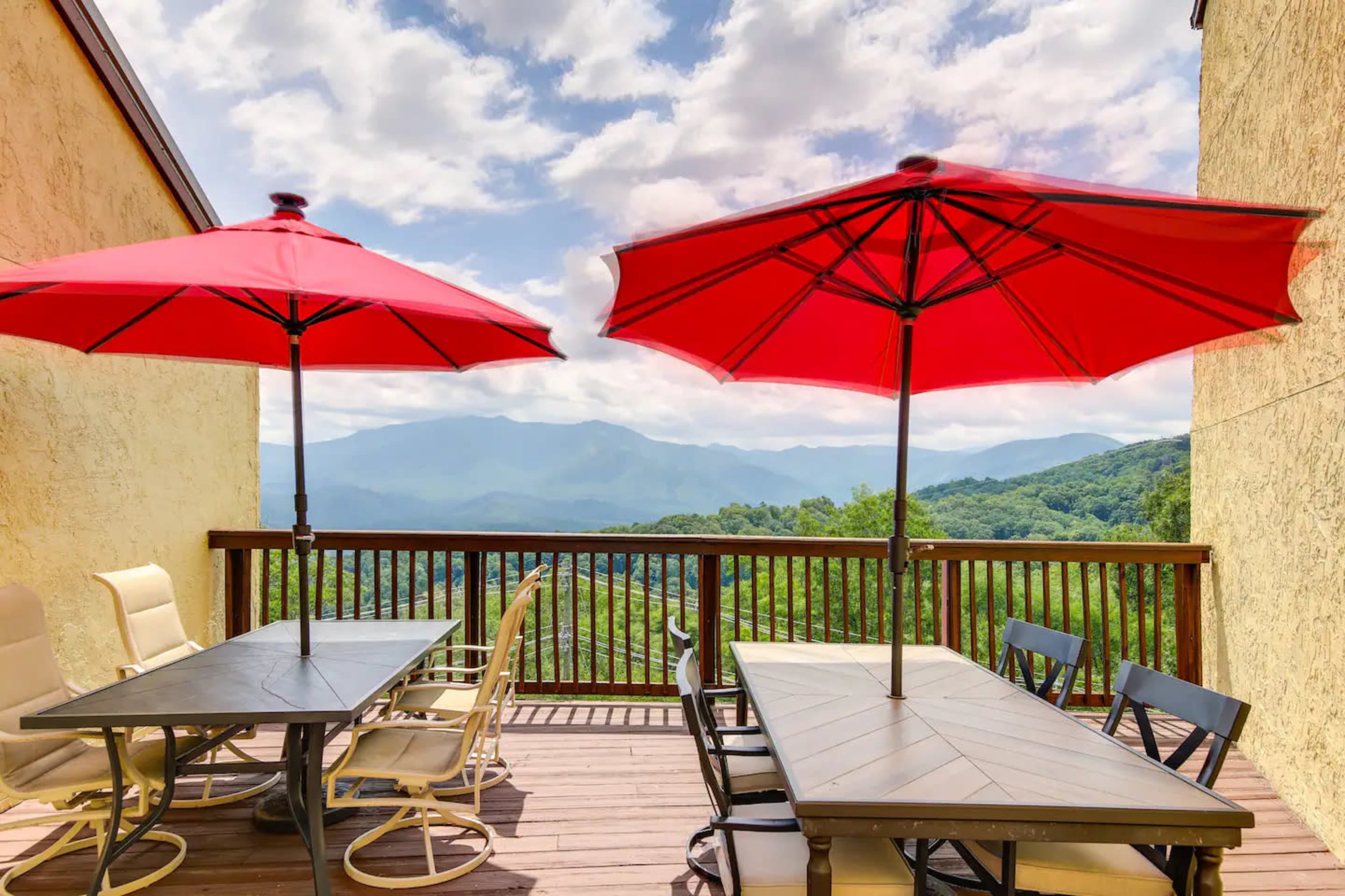 A deck features two red umbrellas over tables, with a mountainous landscape visible in the background.