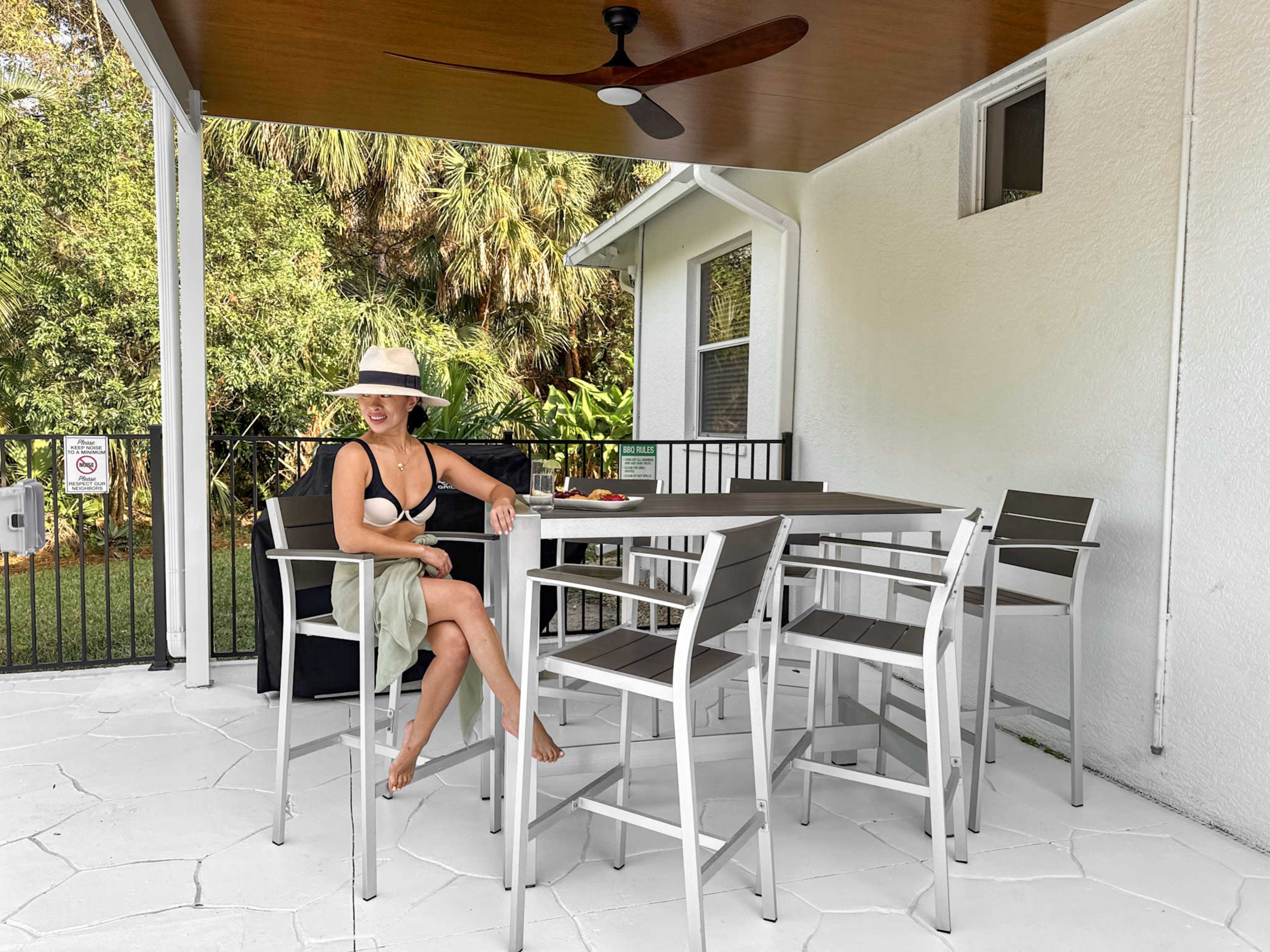 A woman in a black top and hat sits at a table surrounded by metal chairs on a covered patio.