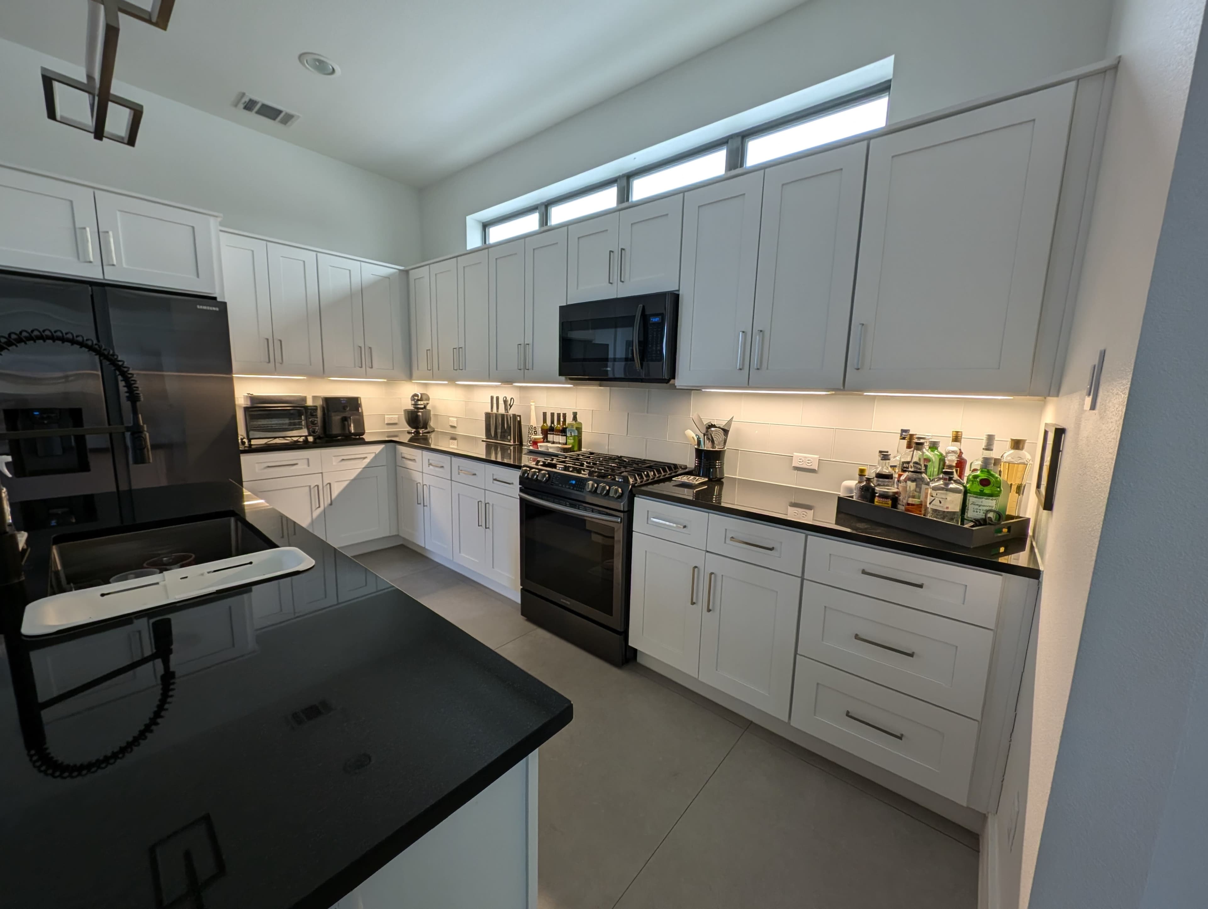 A modern kitchen features white cabinetry, a black countertop, and stainless steel appliances, with a well-organized arrangement of kitchenware and bottles on display.