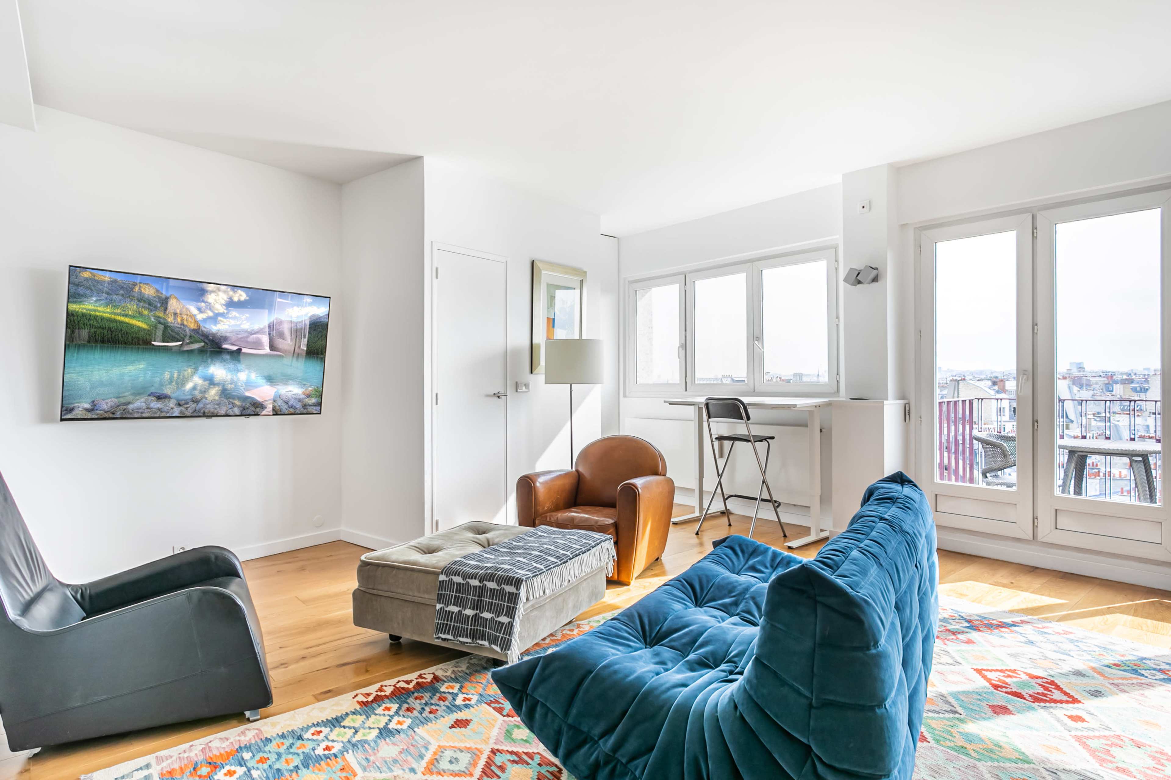 The image shows a modern living room featuring a large television mounted on the wall, a blue upholstered chair, a brown leather armchair, and a small table by the windows with a view.