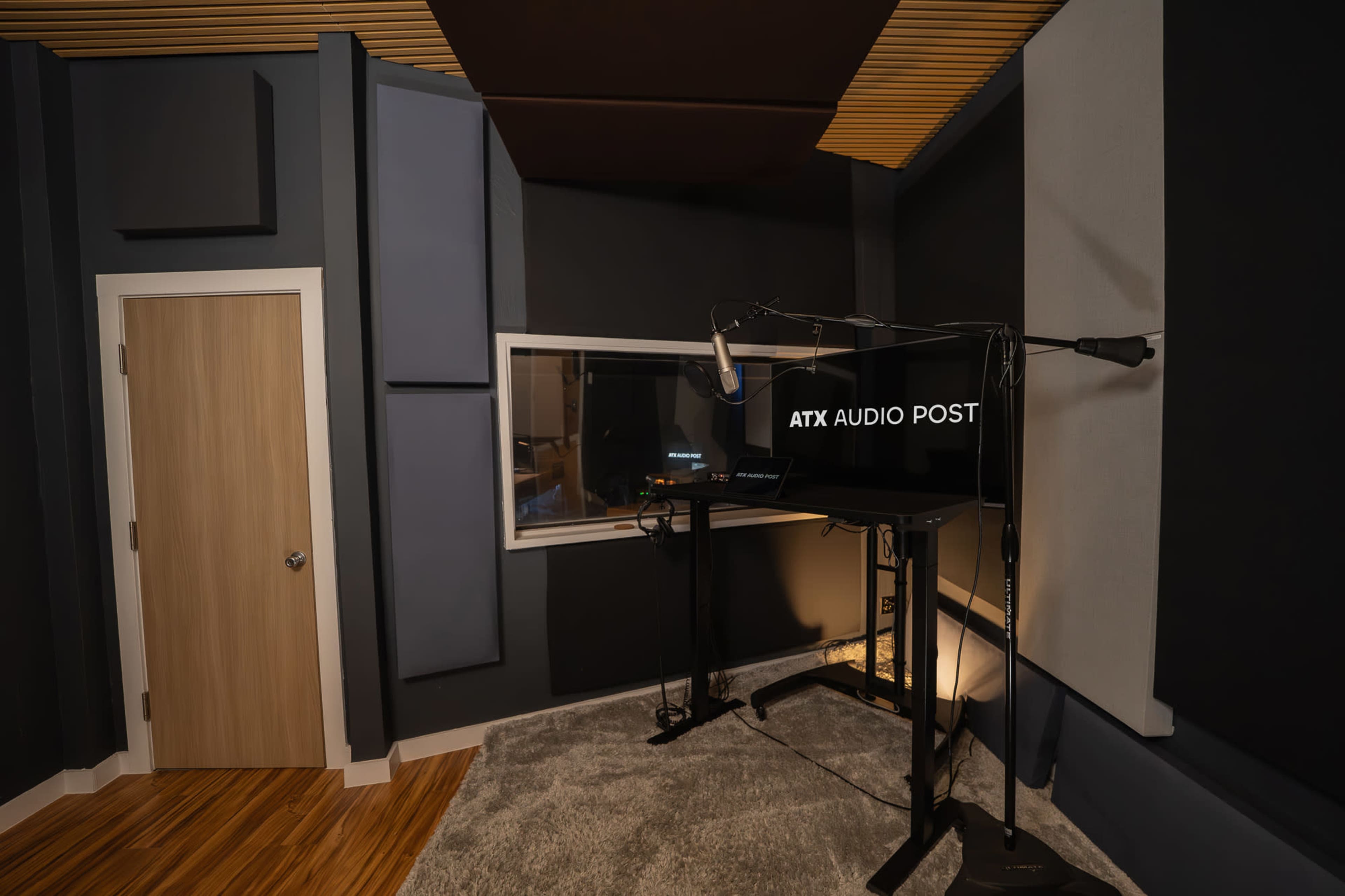 The image shows a soundproof recording studio with acoustic panels, a microphone setup, and a window into an adjacent control room.