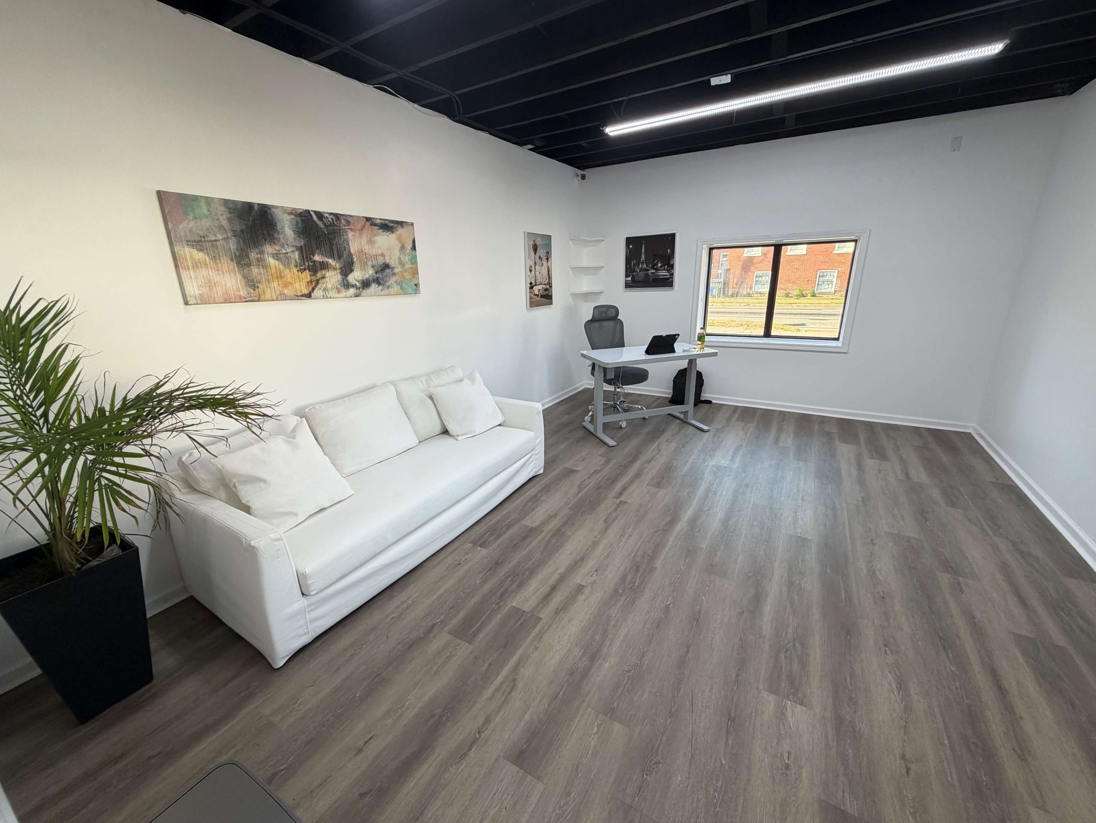 The image shows a minimalist office space with a white sofa, a desk and chair, and a large window allowing natural light to enter.