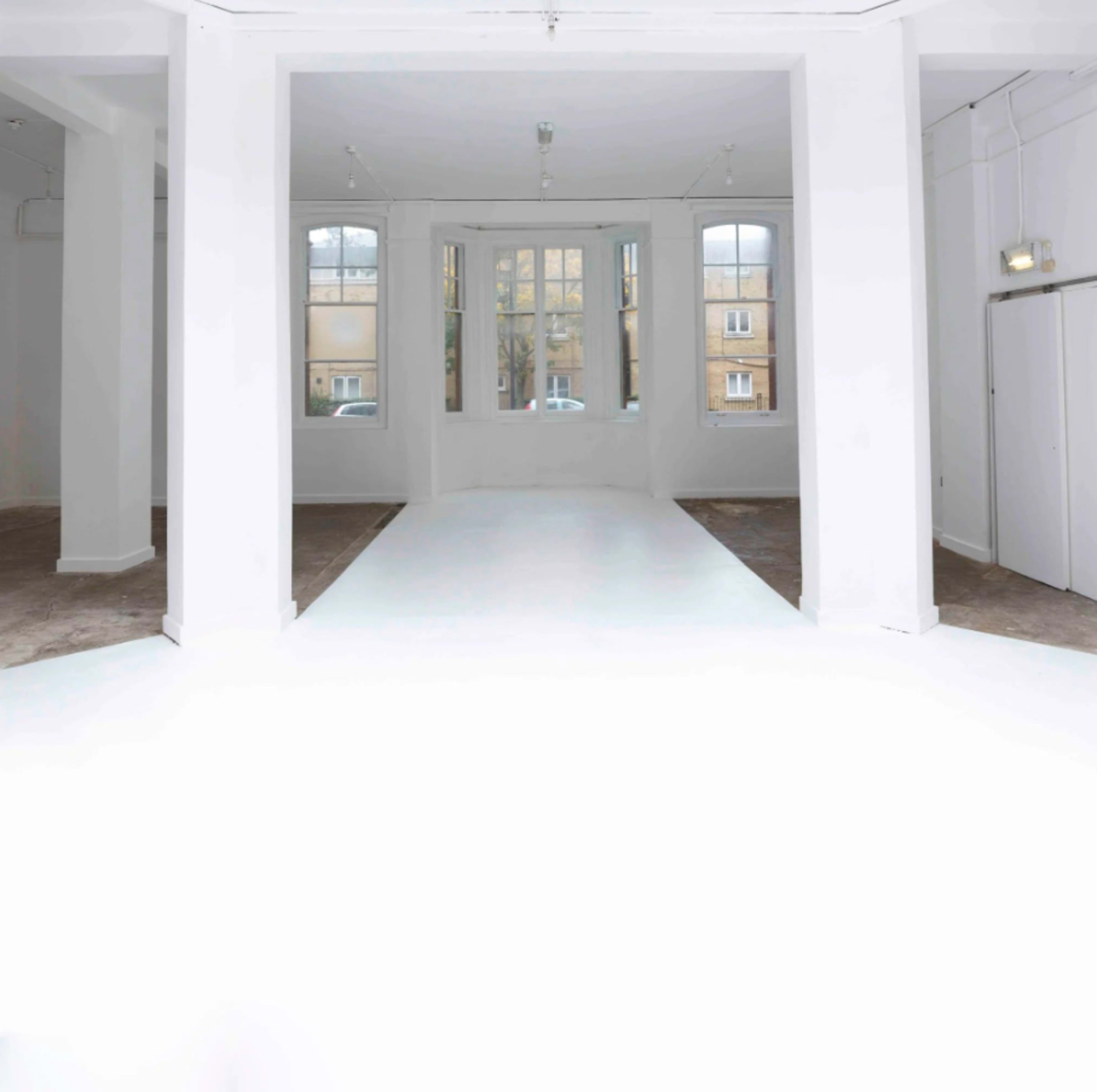The image shows a spacious, empty indoor area with white walls and a light-colored floor, featuring large windows that allow natural light in.