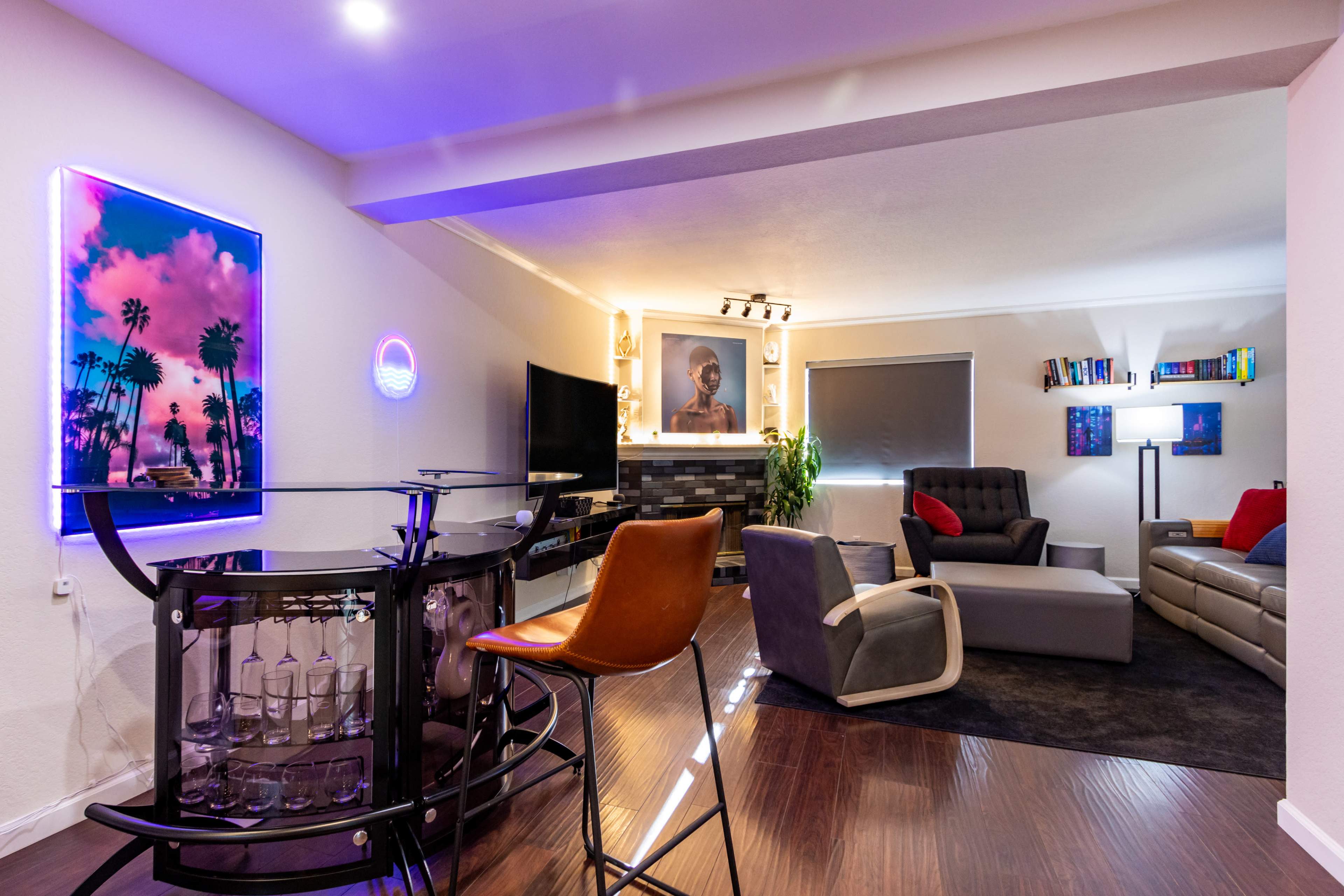 The image shows a modern living room featuring a seating area with a gray sofa, a black armchair, a bar with stools, and a decorative wall with artwork and a neon sign.
