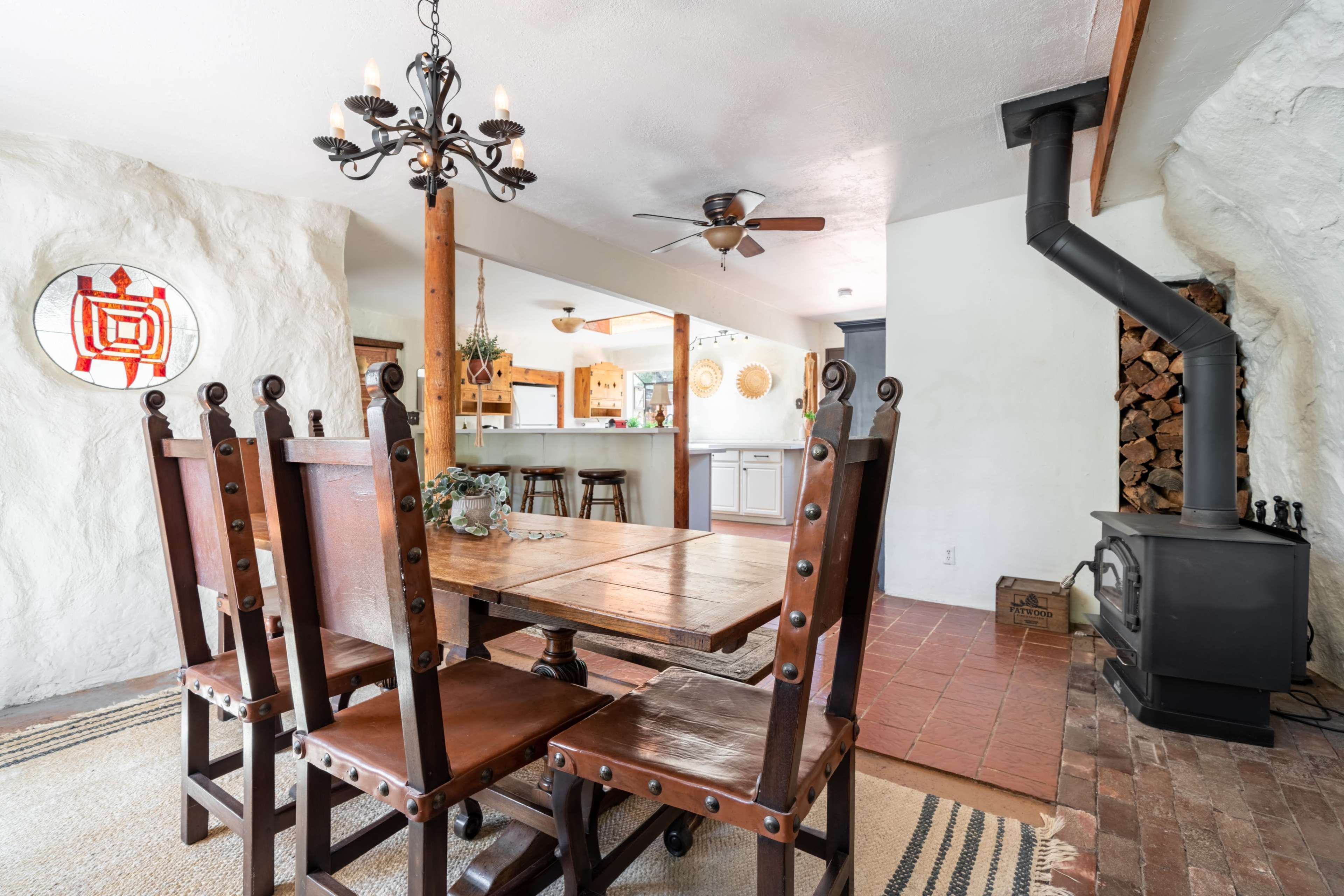 A dining area with a wooden table and chairs, a chandelier overhead, and a stove in the corner, with a view into a kitchen.