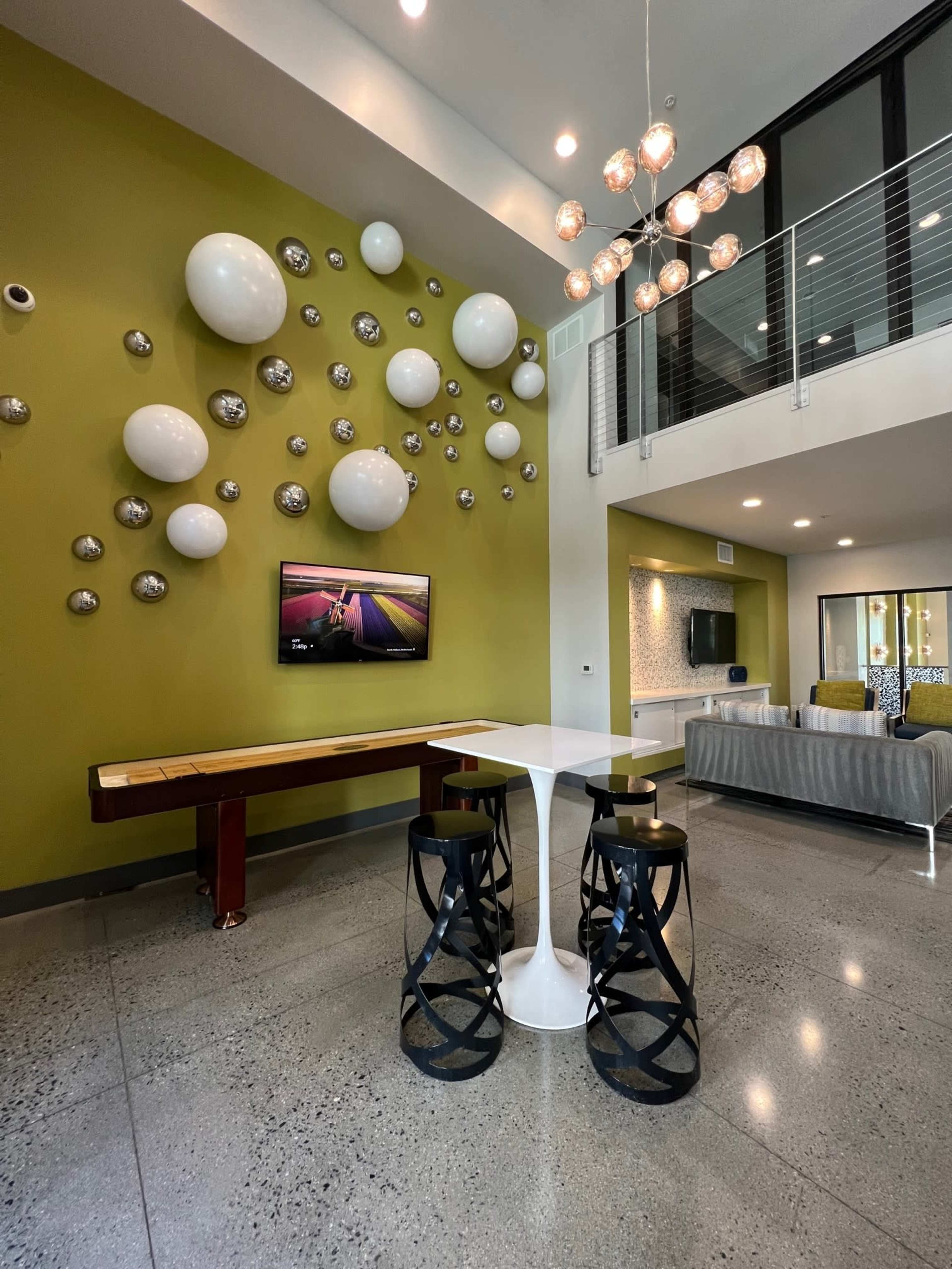 A modern communal lounge area featuring a green wall adorned with round white and silver decorations, a television mounted on the wall, a minimalist white dining table with black stools, and a seating area with a couch in the background.