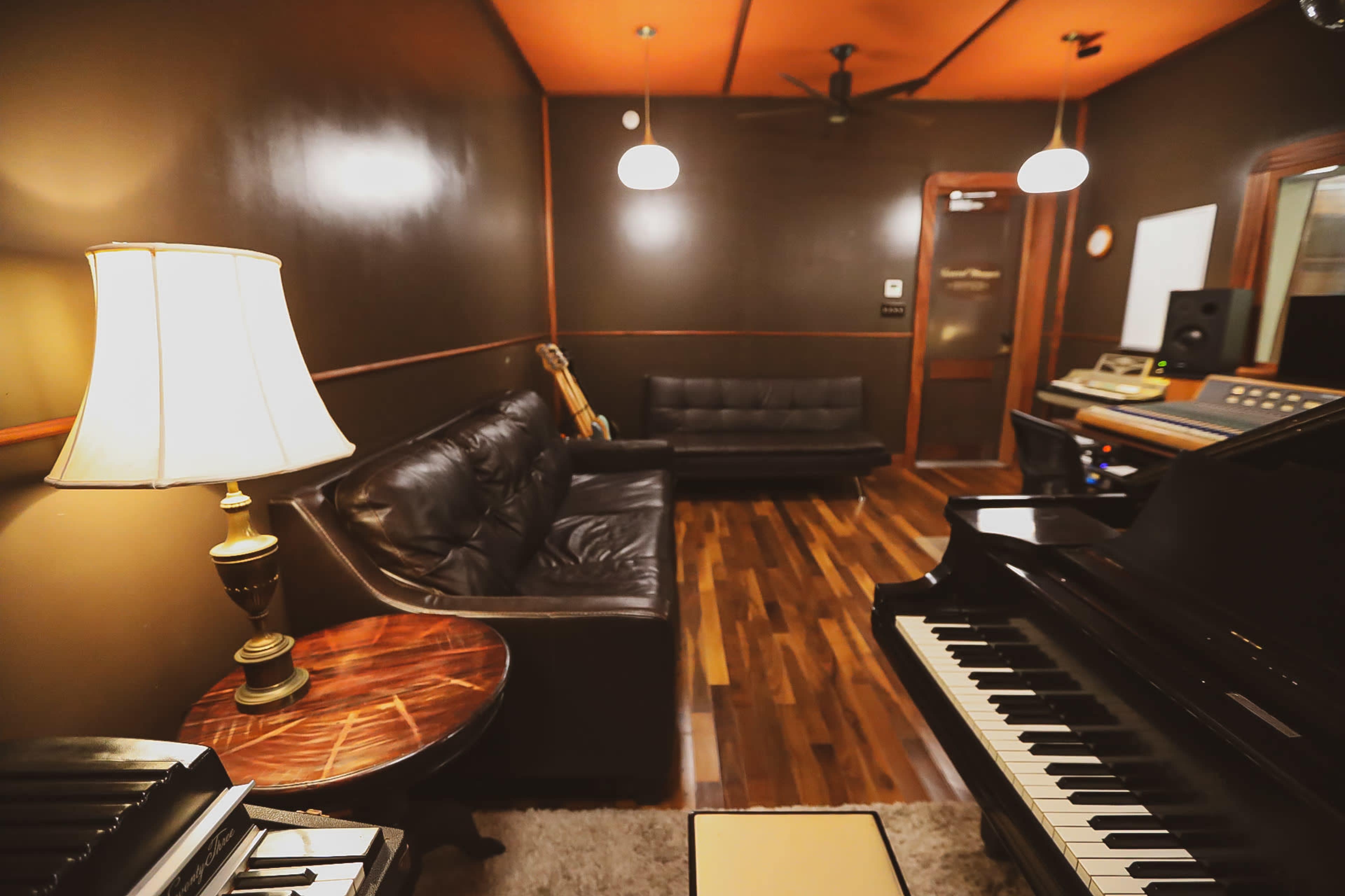 The image shows a small music studio with a grand piano, a couch, and a table under soft lighting.