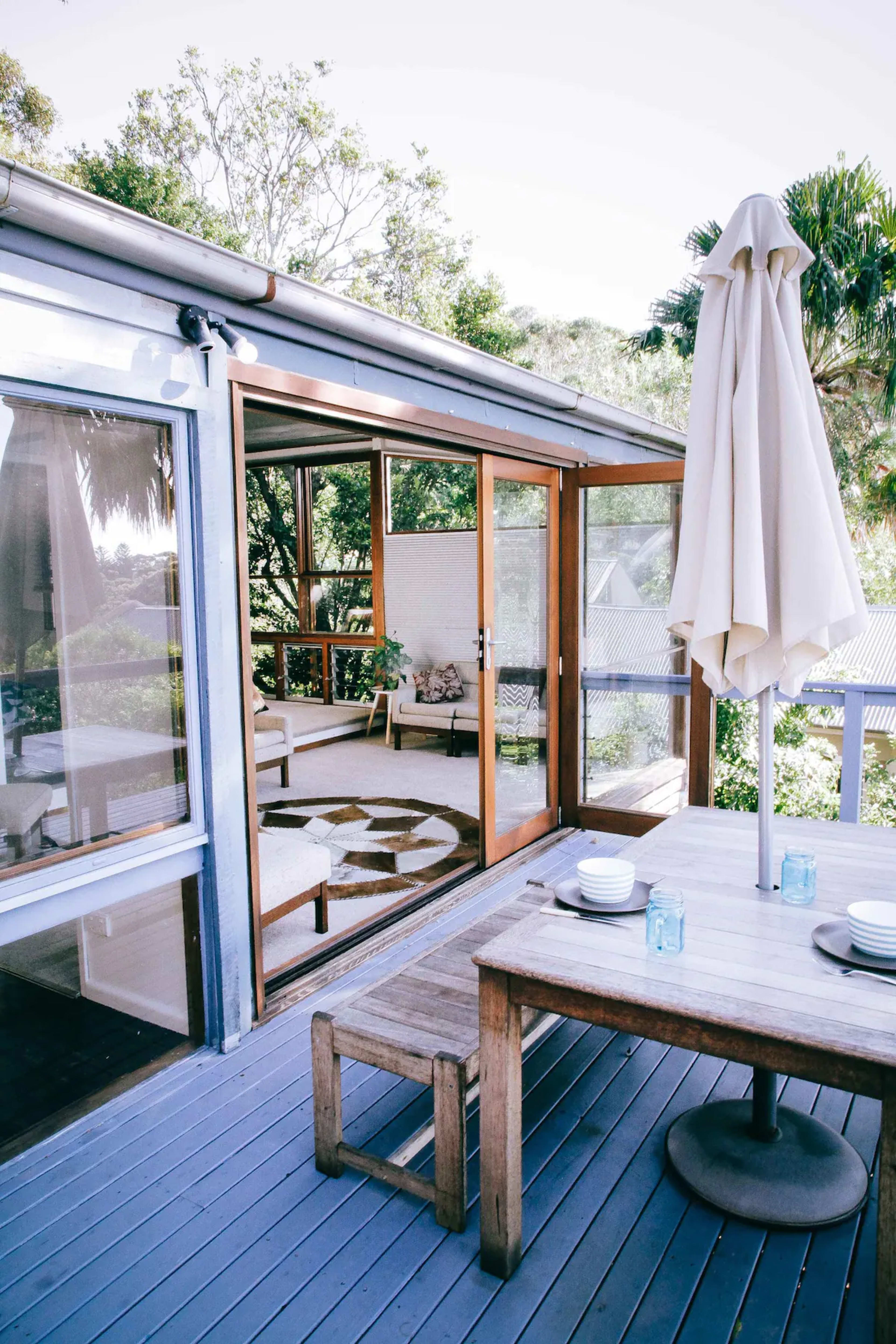 A patio area features a wooden table and chairs with a large umbrella overlooking a light-filled interior space through glass sliding doors.