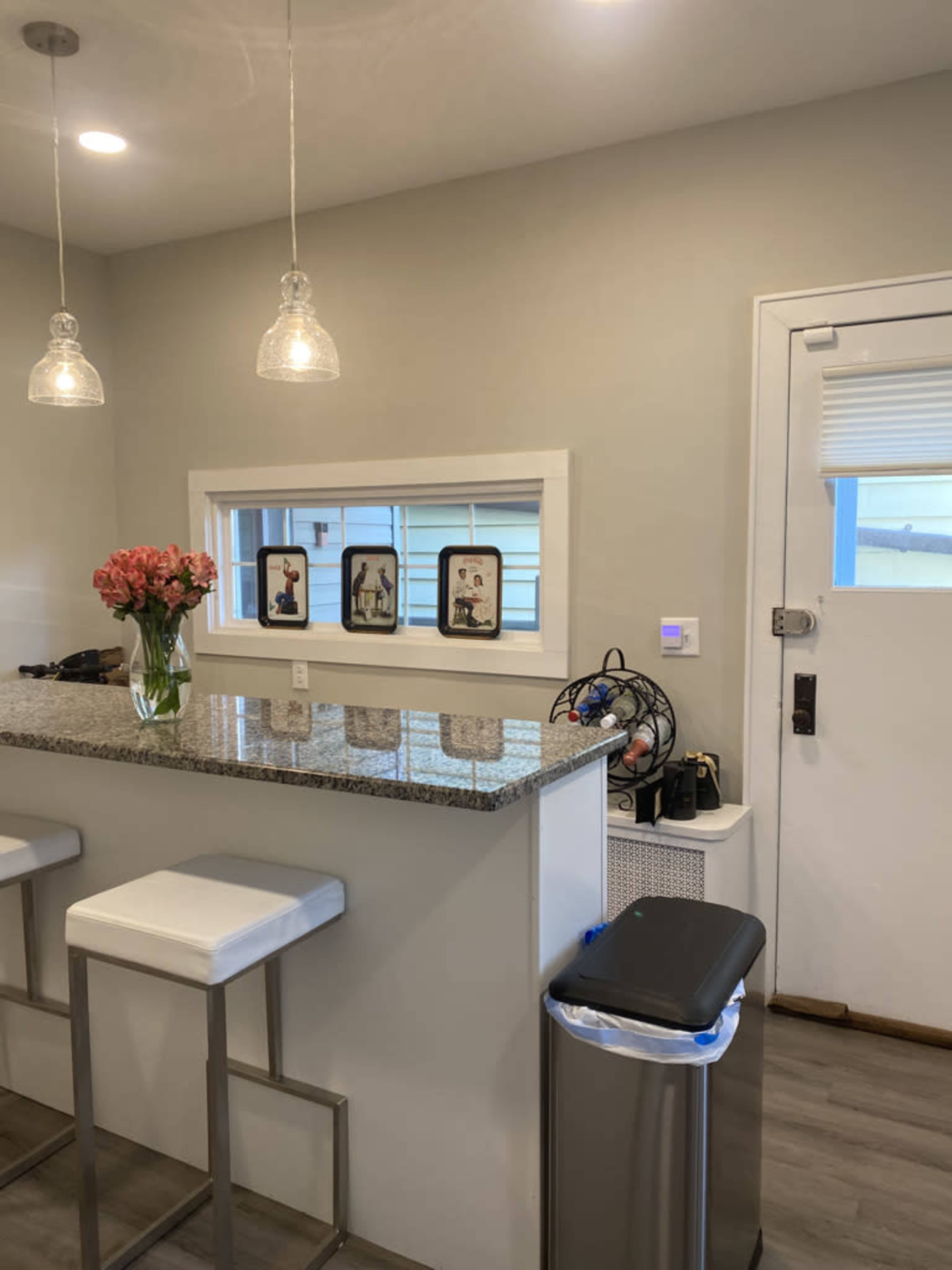 A modern kitchen features a granite countertop with two bar stools, a vase of flowers, and a window displaying framed pictures.