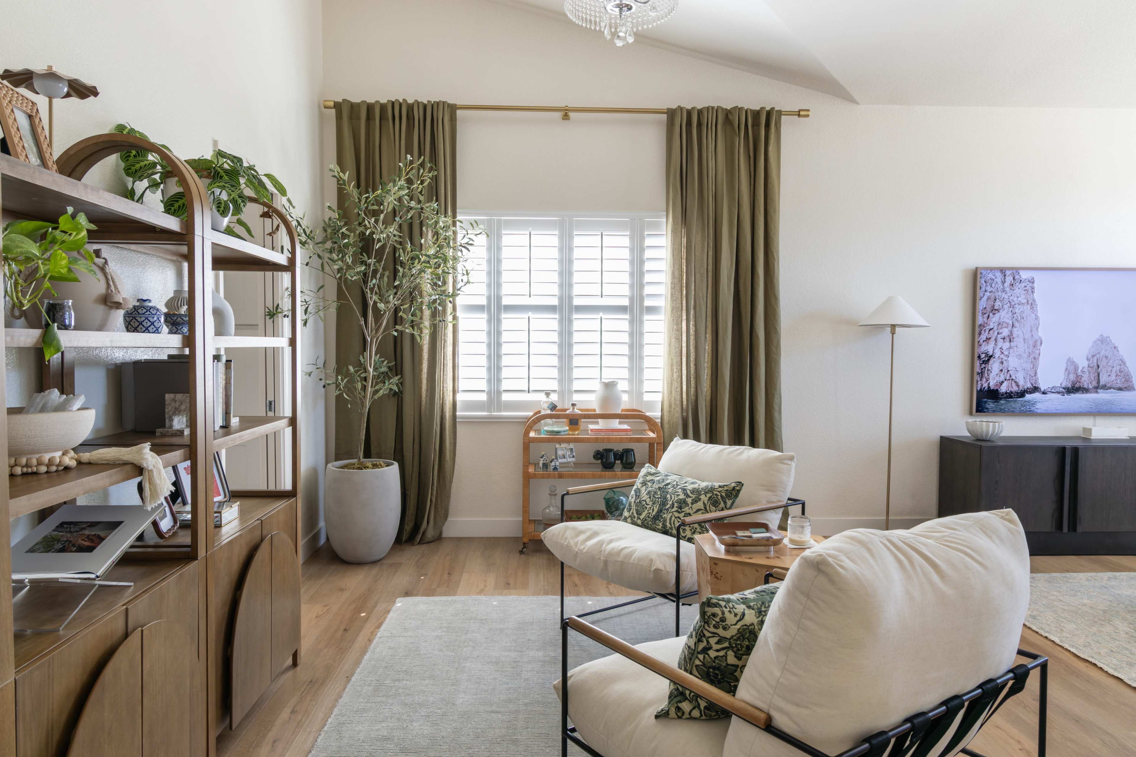 The image shows a modern living room featuring a seating area with two chairs, a large window with curtains, a bookshelf, and a television mounted on the wall.