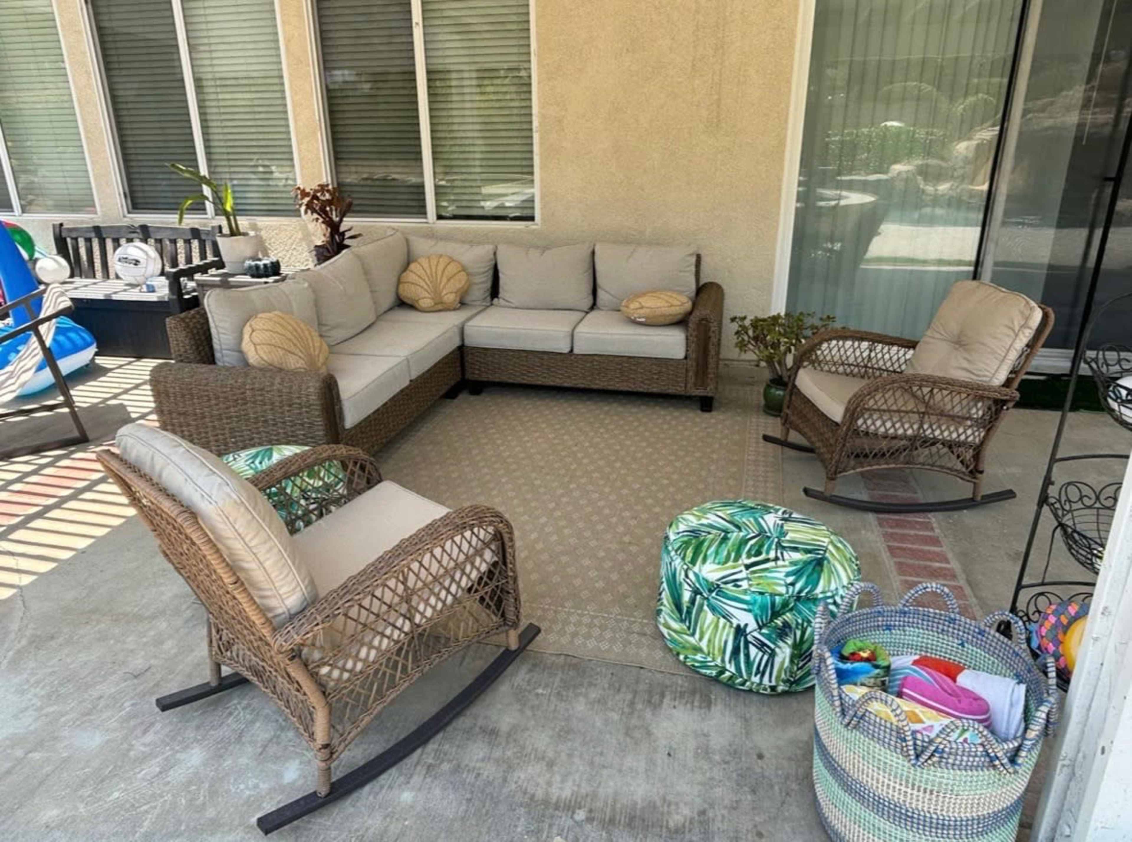 The image shows a covered patio area with a sectional sofa, two rocking chairs, and various cushions and decor items scattered around.