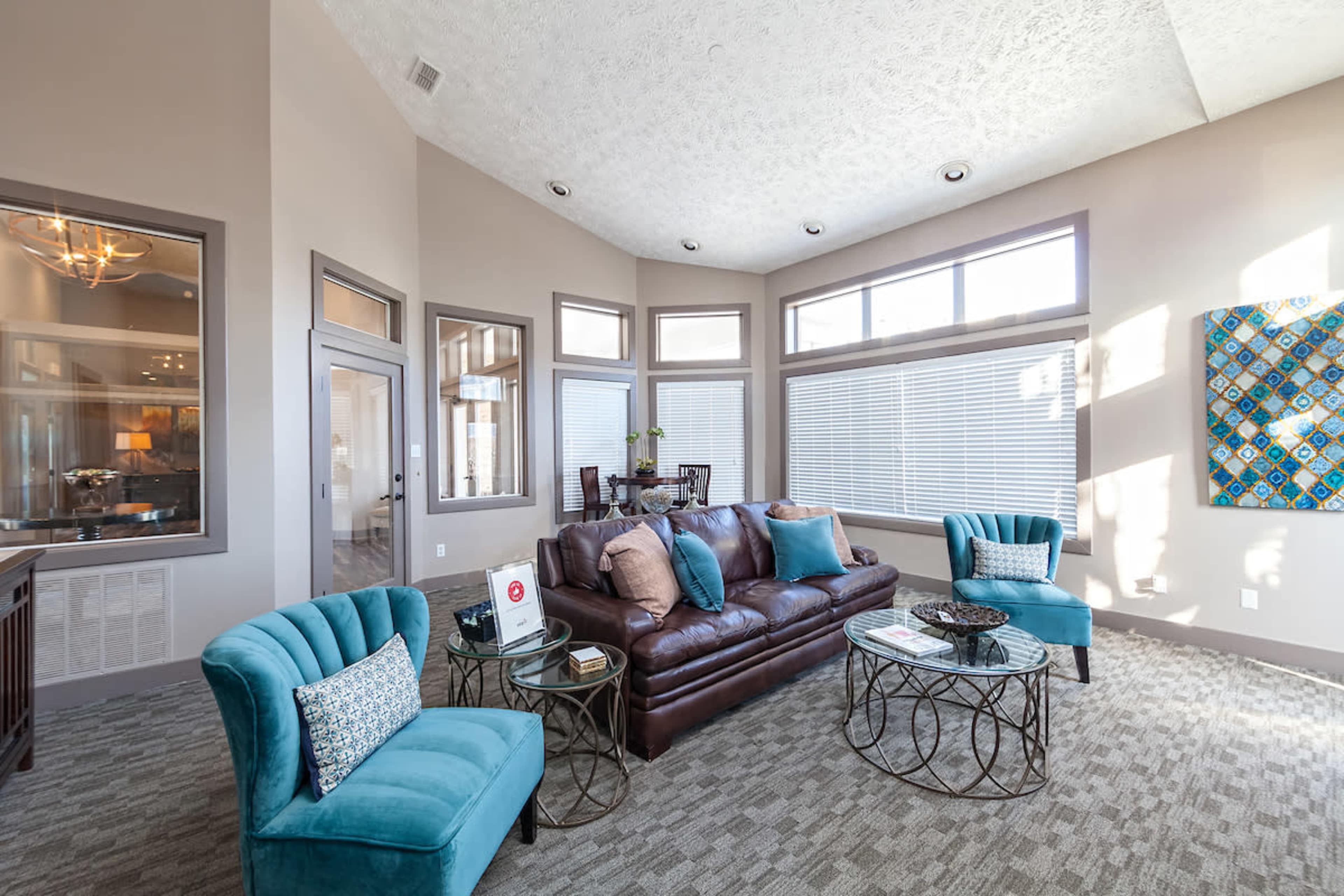 A modern living room with a brown leather sofa, two turquoise chairs, and large windows allowing natural light to fill the space.