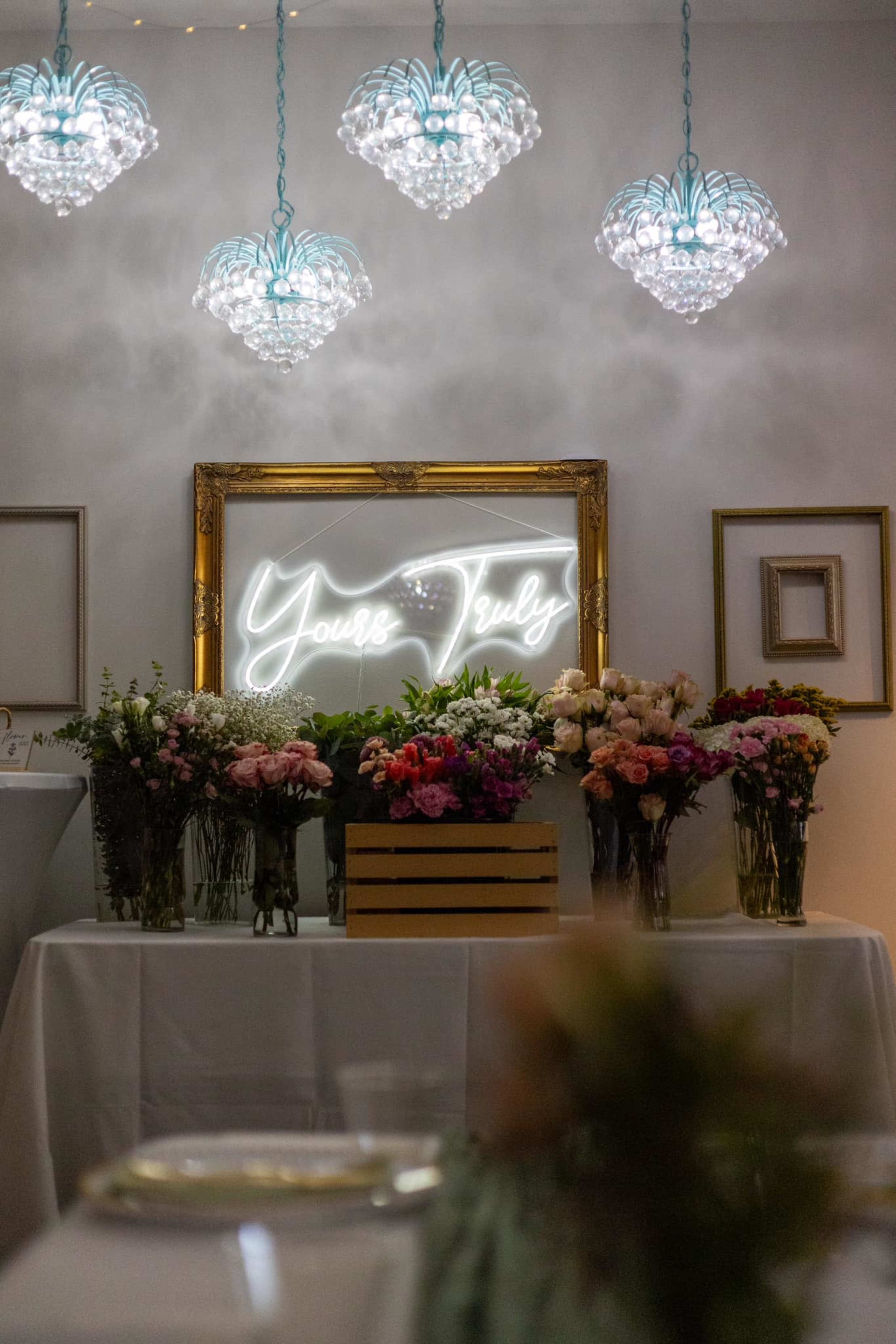 A beautifully arranged table features various flower arrangements and a neon sign saying "Yours Truly" framed by ornate lighting fixtures.