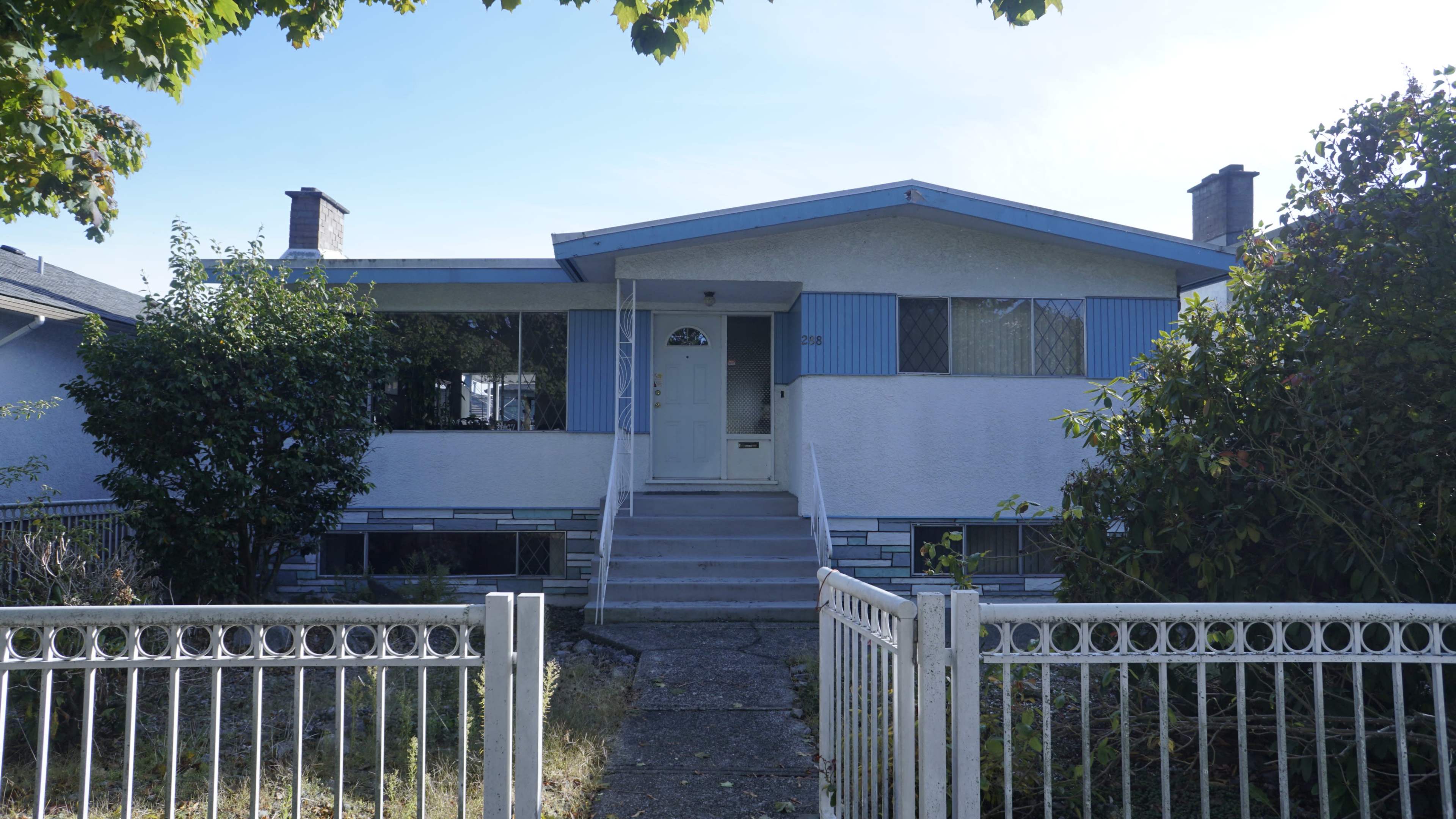 The image shows a beige-colored, single-story house with blue accents and a front porch, surrounded by overgrown shrubs and a white fence.