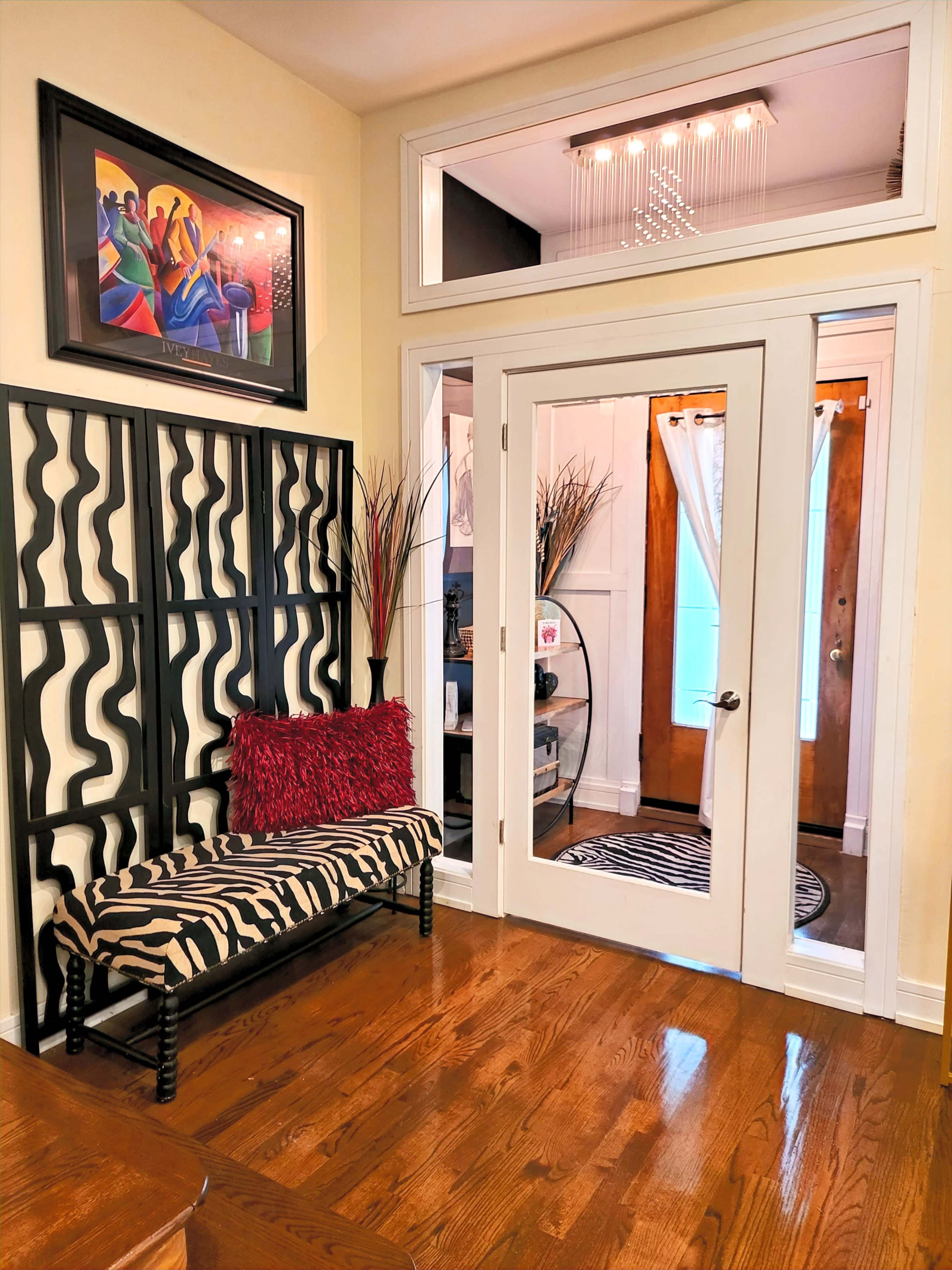 A stylish entryway featuring a black and white zebra-patterned bench, a decorative room divider, and a glass door leading to a bright hallway with a chandelier and artwork on the walls.