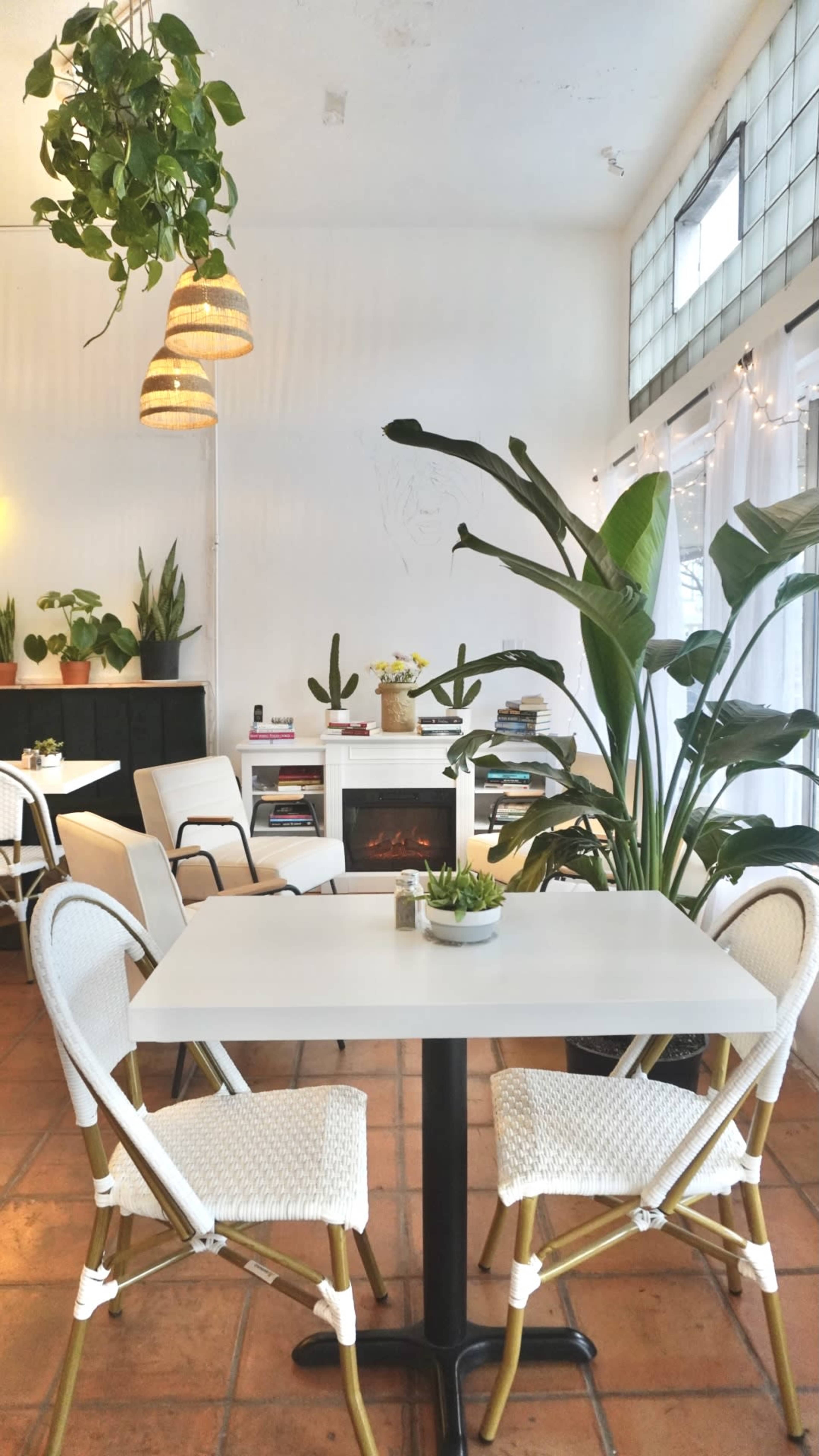 A cozy café interior with a white table and chairs, surrounded by potted plants and warm lighting fixtures.