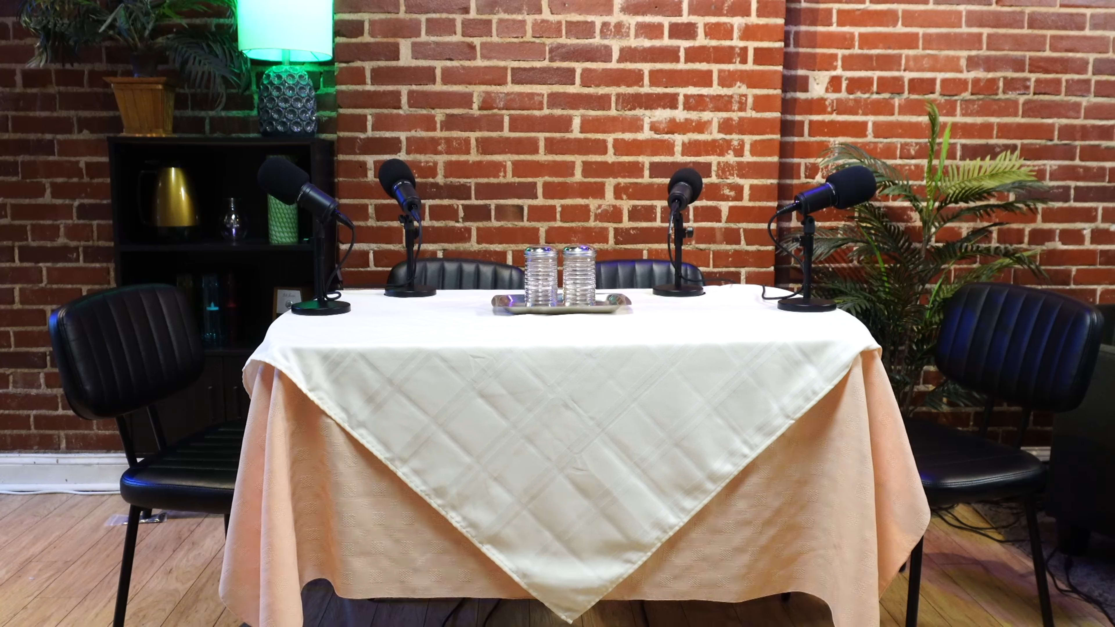 A table covered with a cream-colored tablecloth is set between four microphones on stands, against a backdrop of exposed brick walls and a green-lit shelf.