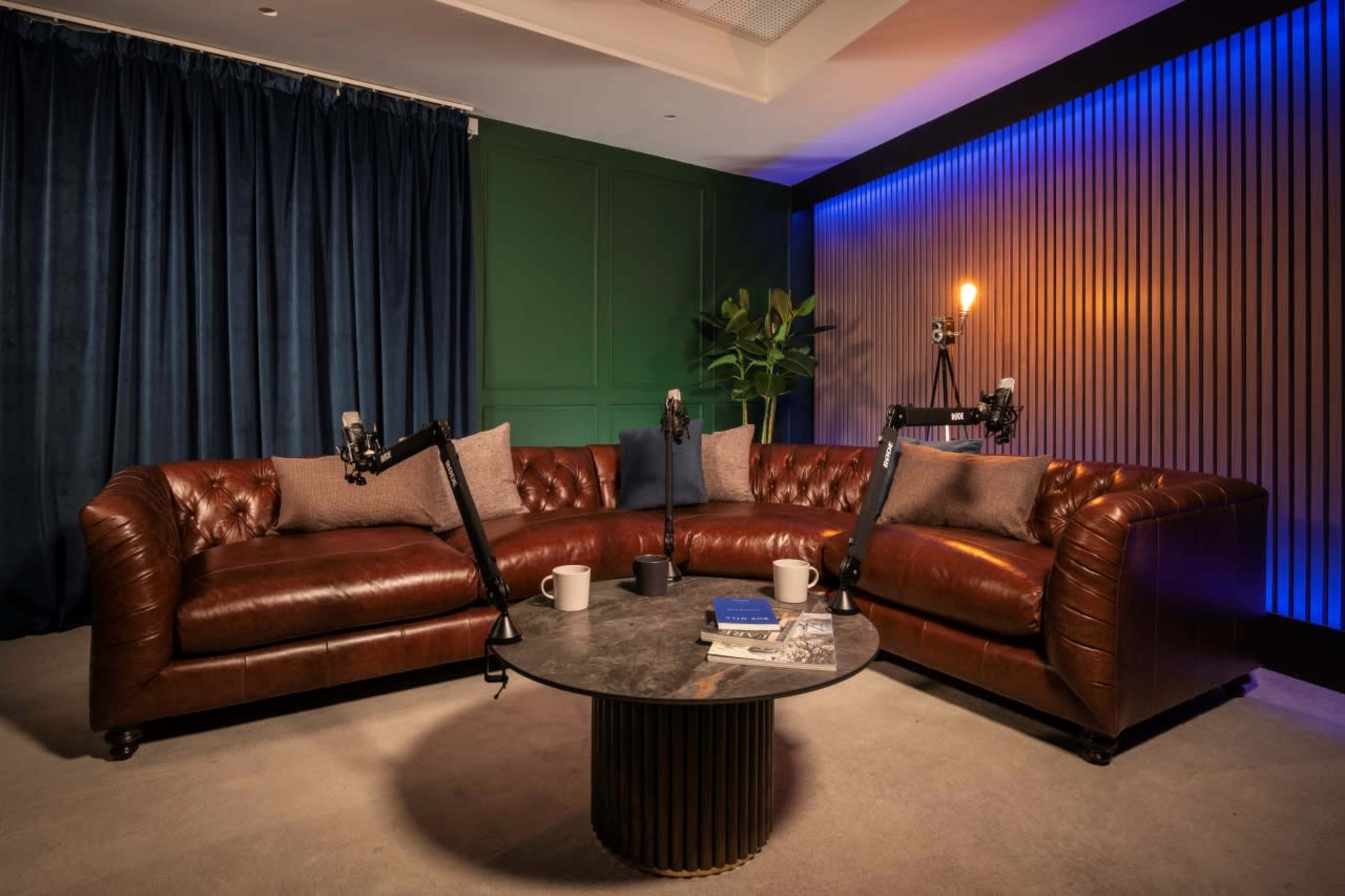 A cozy, modern room with a brown leather sofa, a circular coffee table, and two microphones on stands, surrounded by dark curtains and colorful wall lighting.