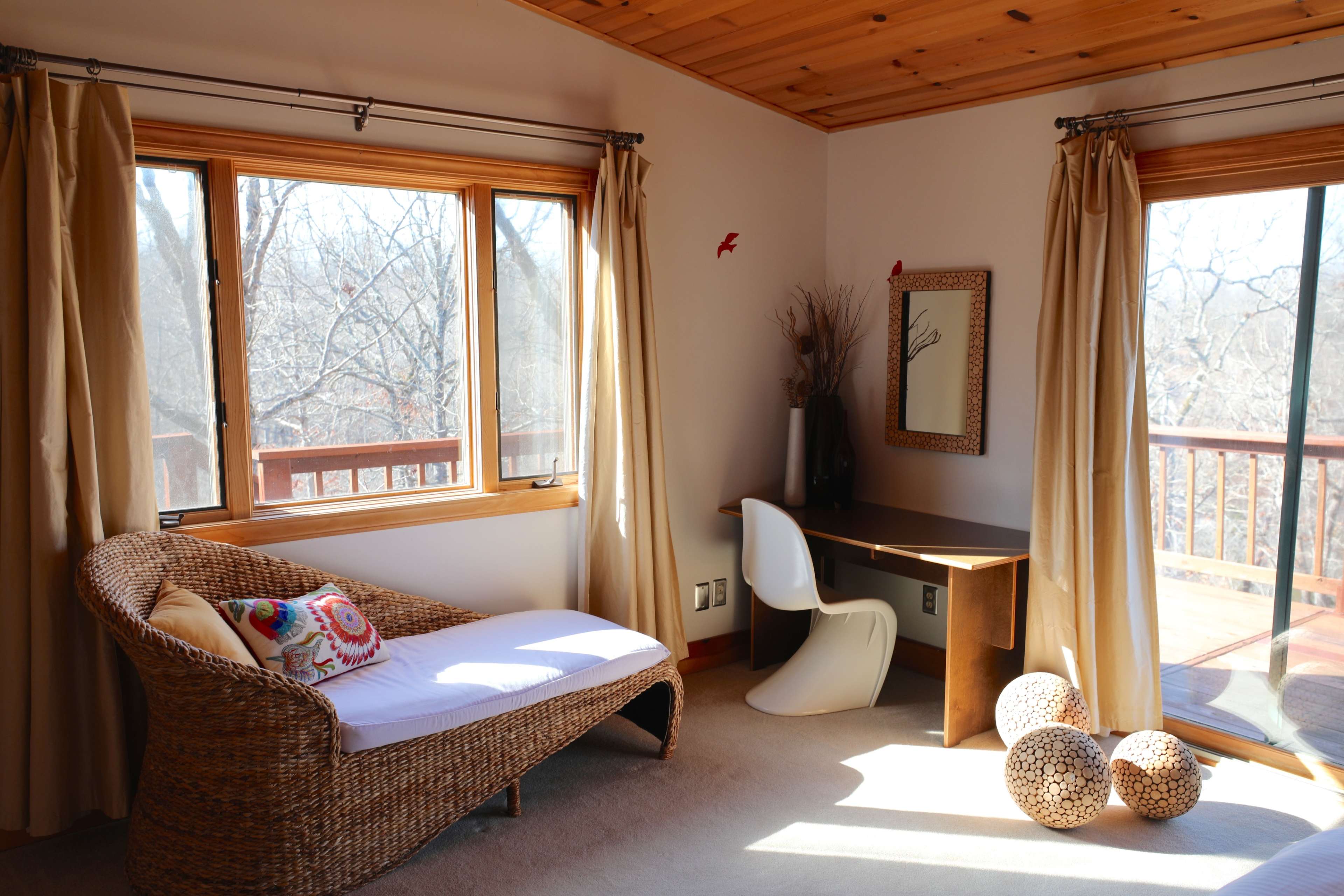 A cozy room with a wicker lounge chair, a modern white desk, and large windows offering a view of the outdoors.