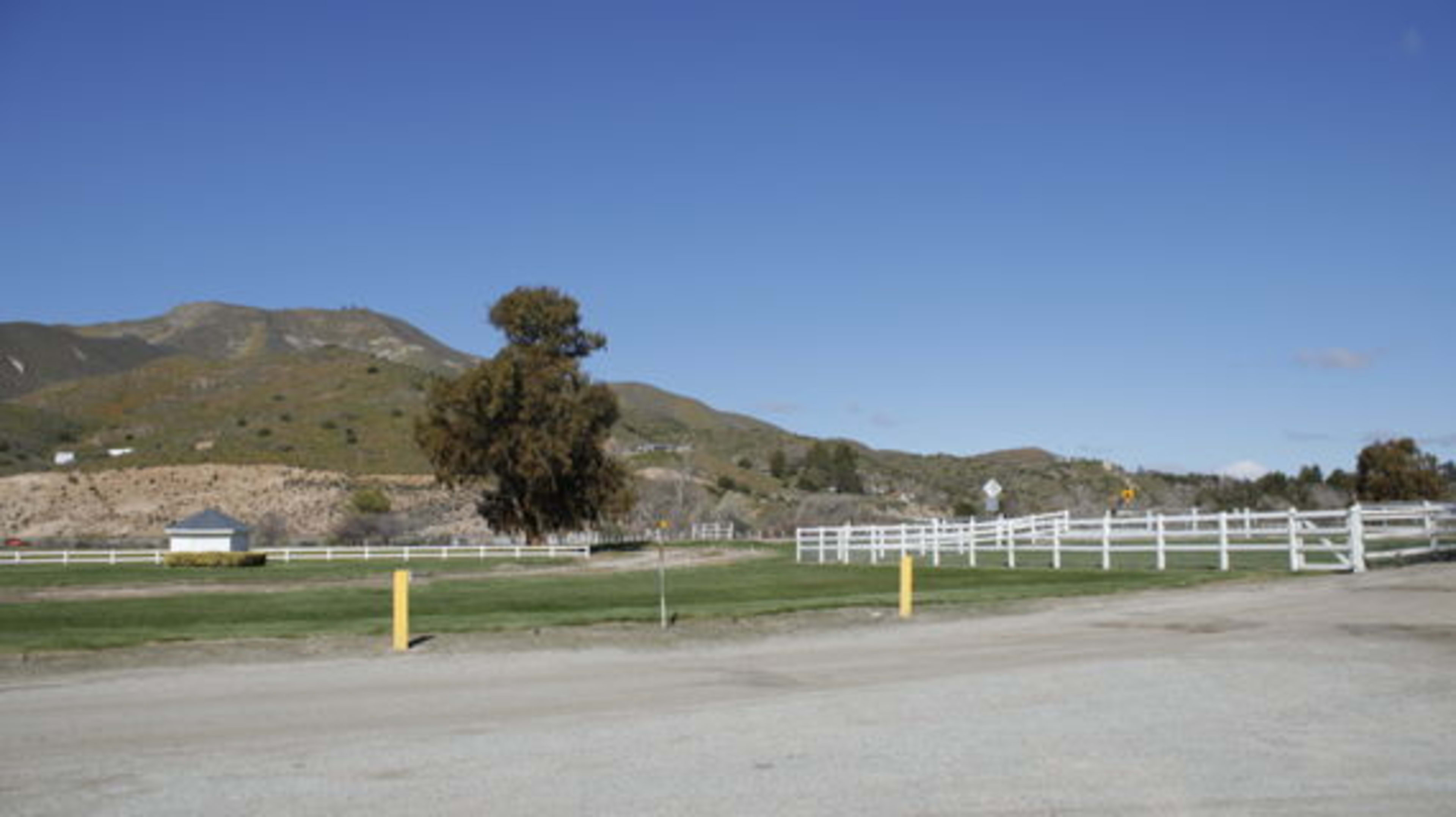 A rural landscape with green grass, a dirt road, a white fence, and a mountainous background under a clear blue sky.