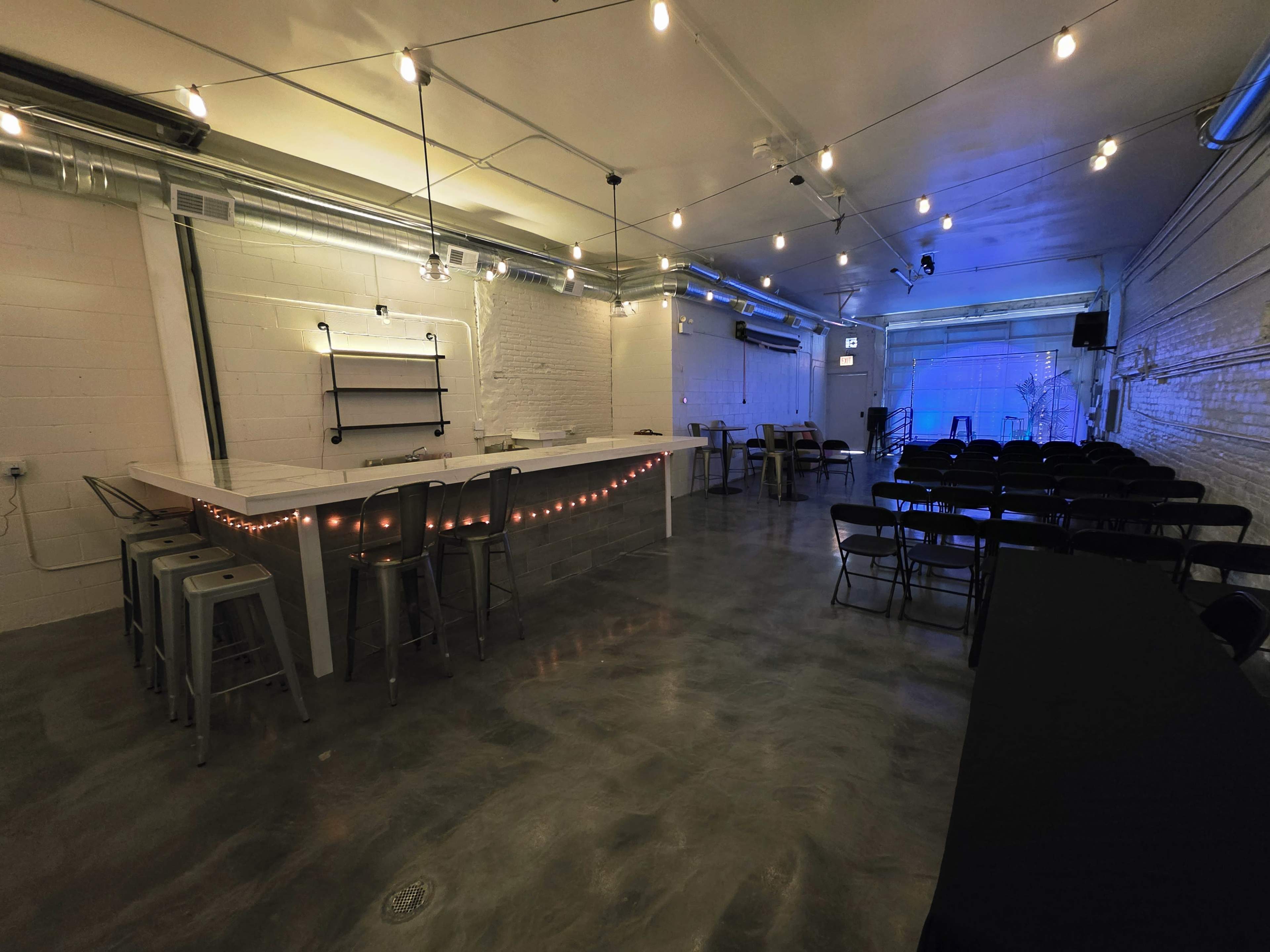 The image shows a spacious, minimalist event room featuring a bar area with string lights, rows of black chairs, and subtle blue lighting on the walls.