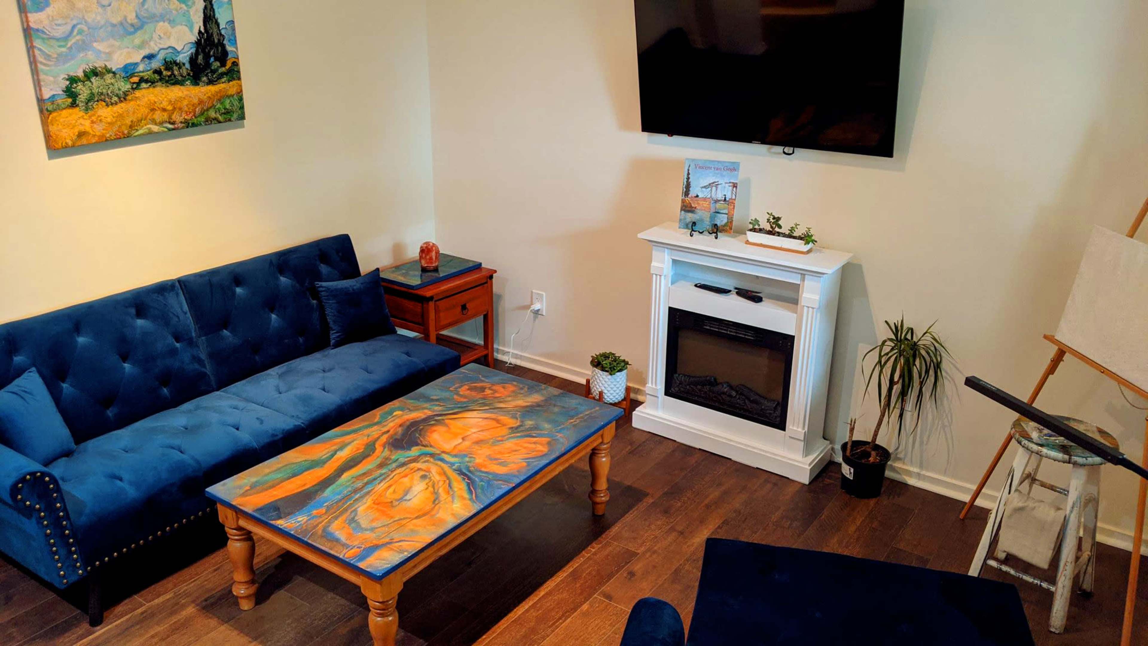The image shows a living room featuring a blue tufted couch, a wooden coffee table with abstract artwork, and a white fireplace beneath a wall-mounted television.