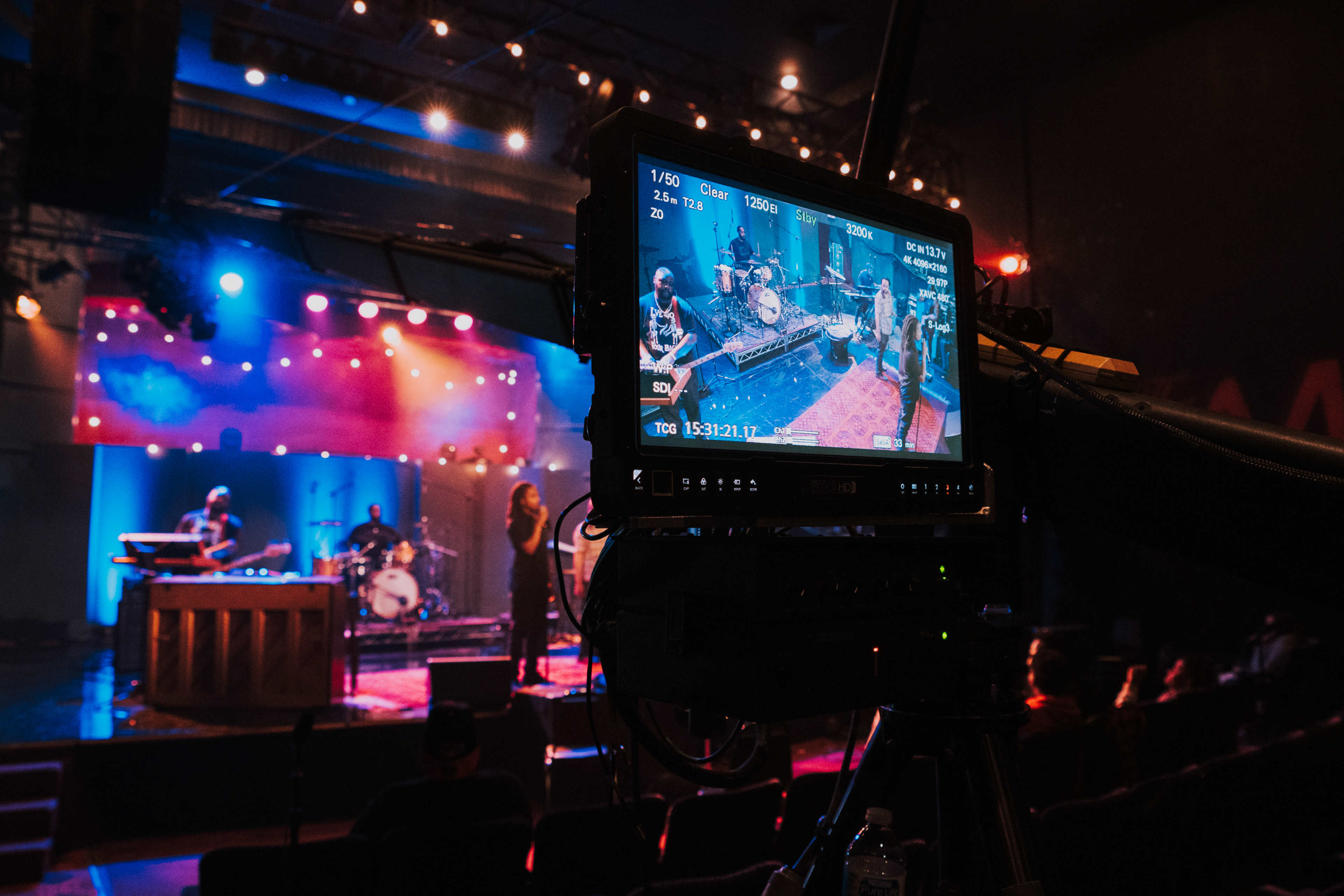 A video camera captures a band performing on stage with colorful lights in a theater setting.