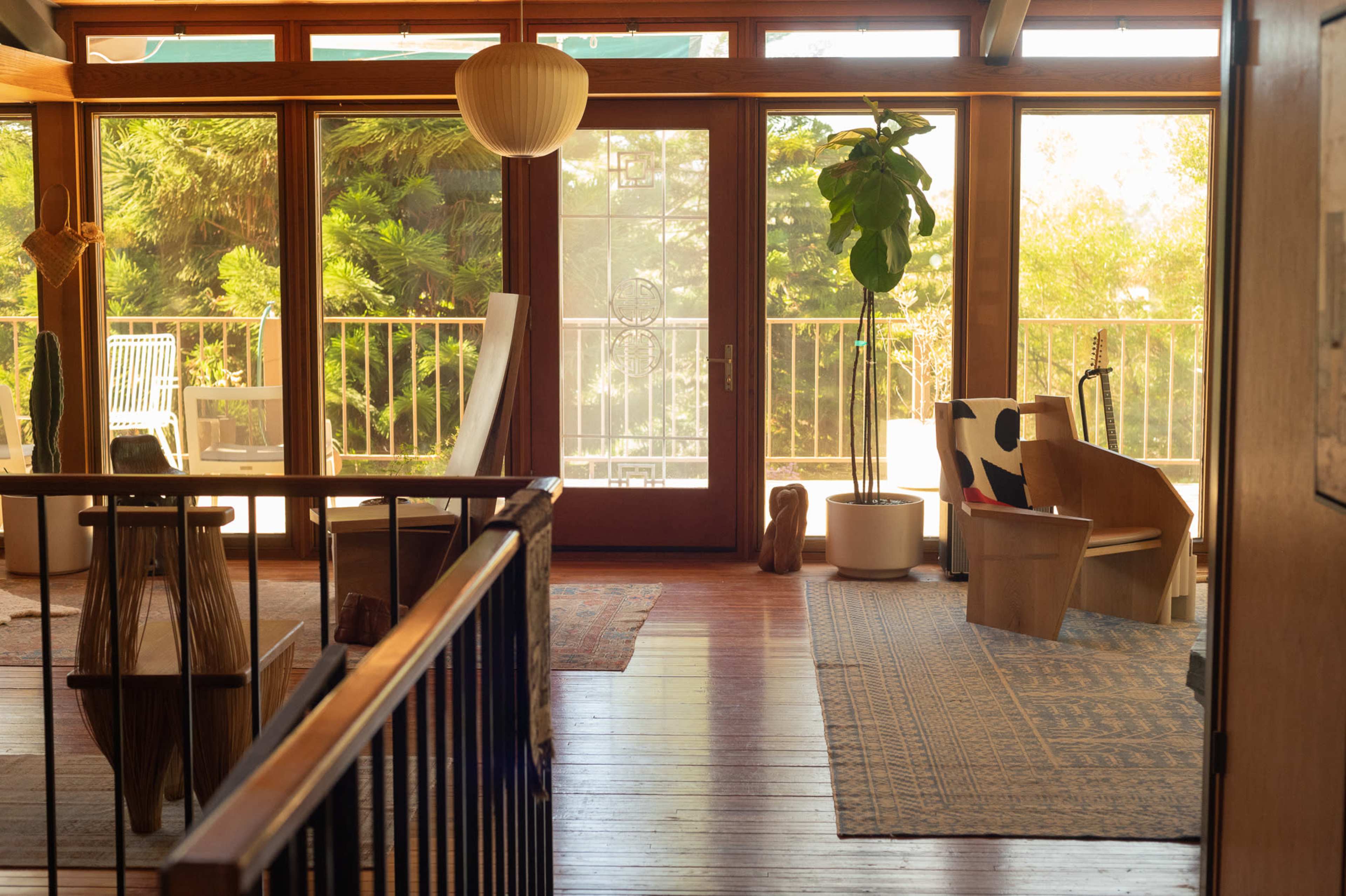 A spacious room features large windows with wooden frames, a potted plant, and various seating arrangements, including a chair and a sculptural decoration.