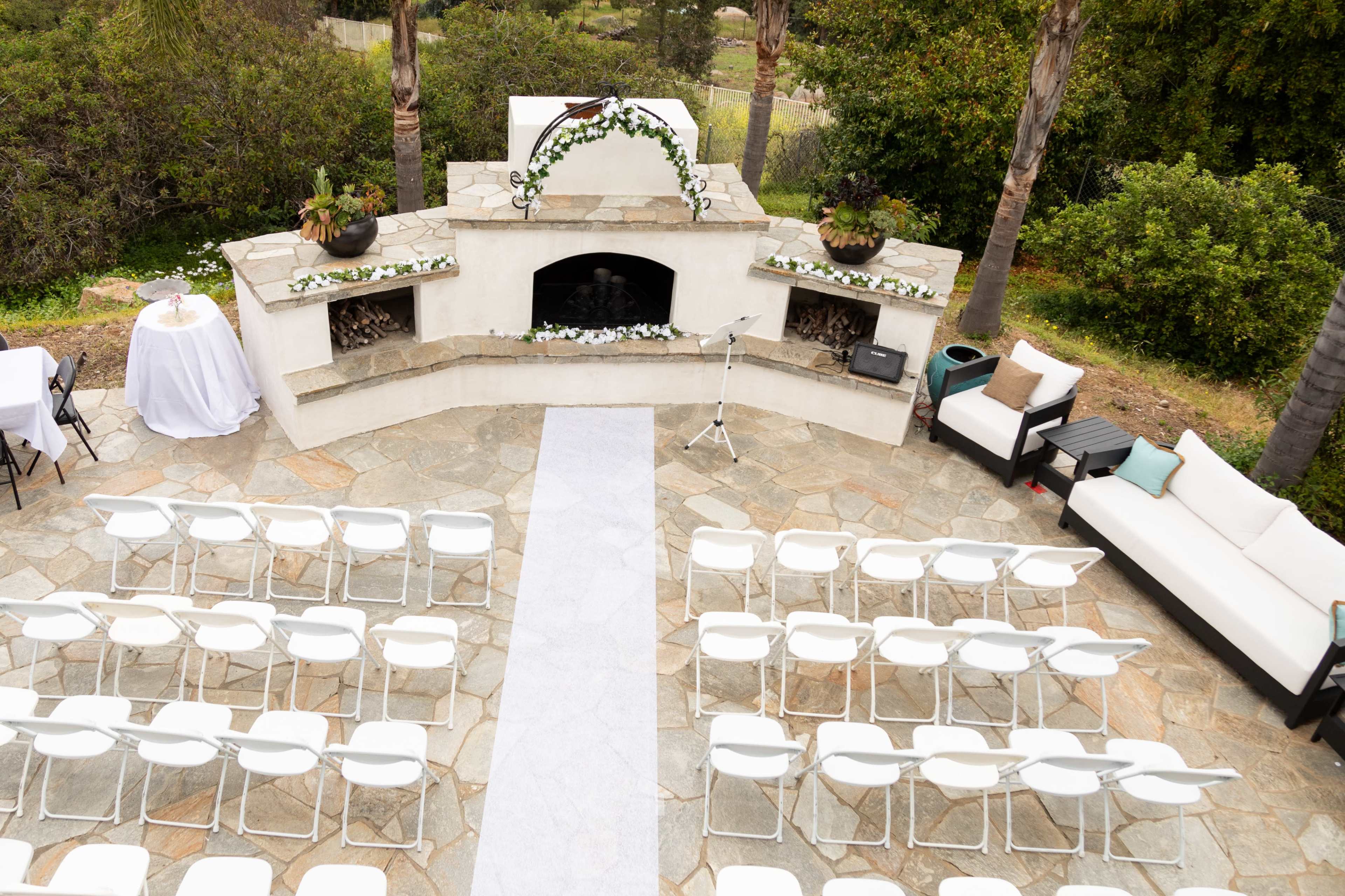 An outdoor wedding setup features white chairs arranged in rows facing a stone fireplace adorned with greenery and floral decorations.