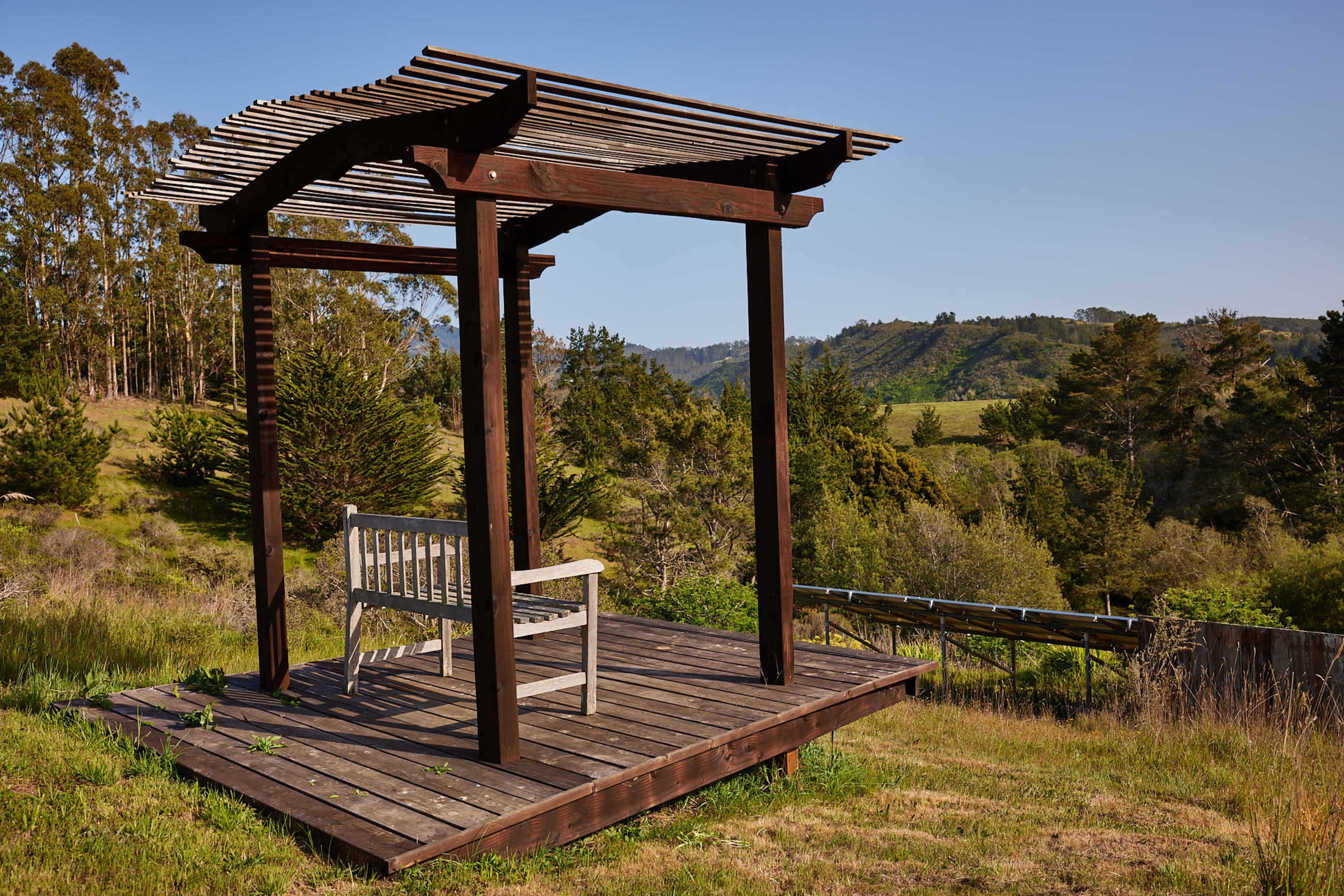 A wooden gazebo with a bench overlooks a grassy hillside and trees.